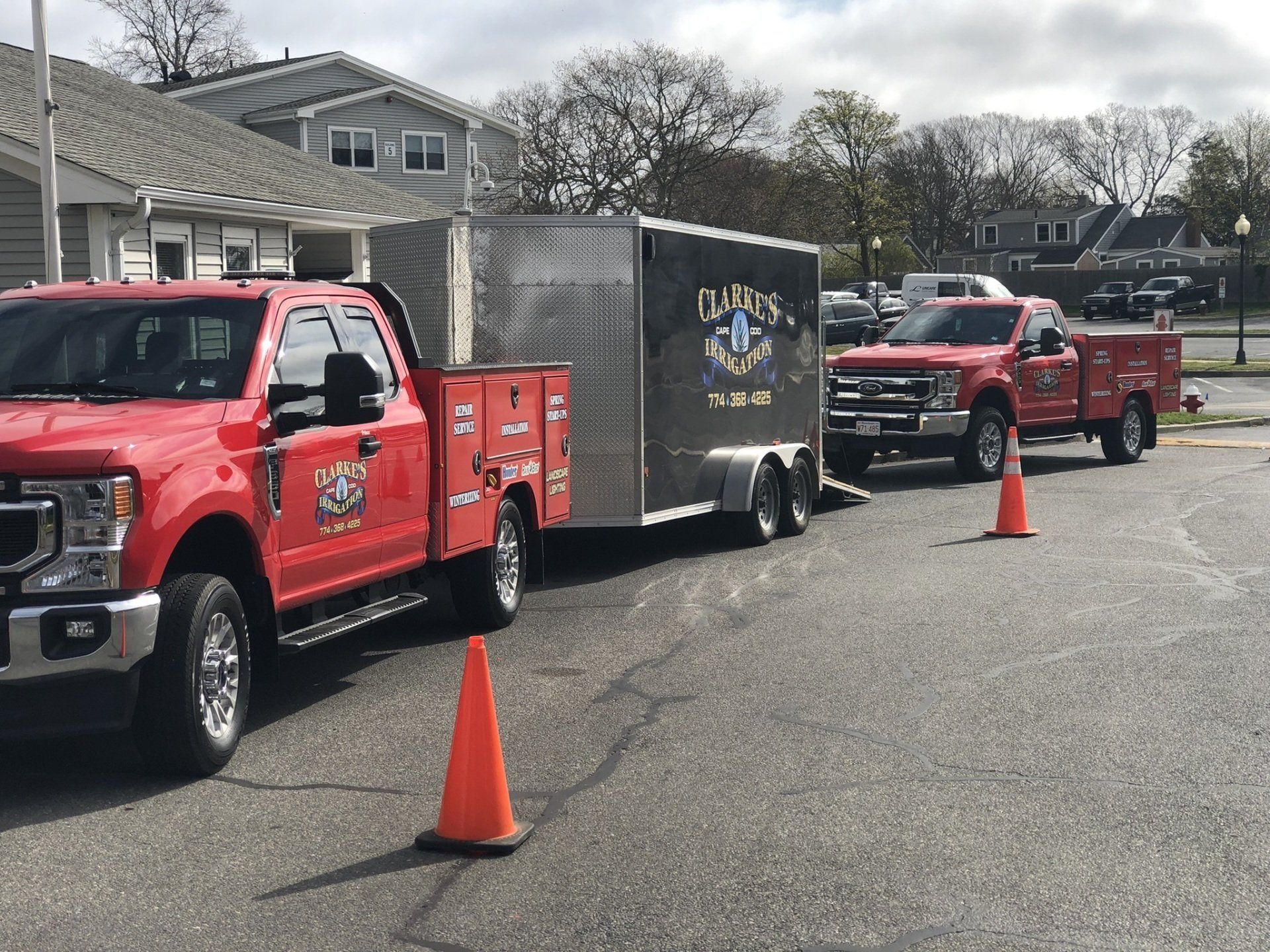 Two red utility trucks parked in a lot, with one towing a dark trailer and orange traffic cones placed nearby.
