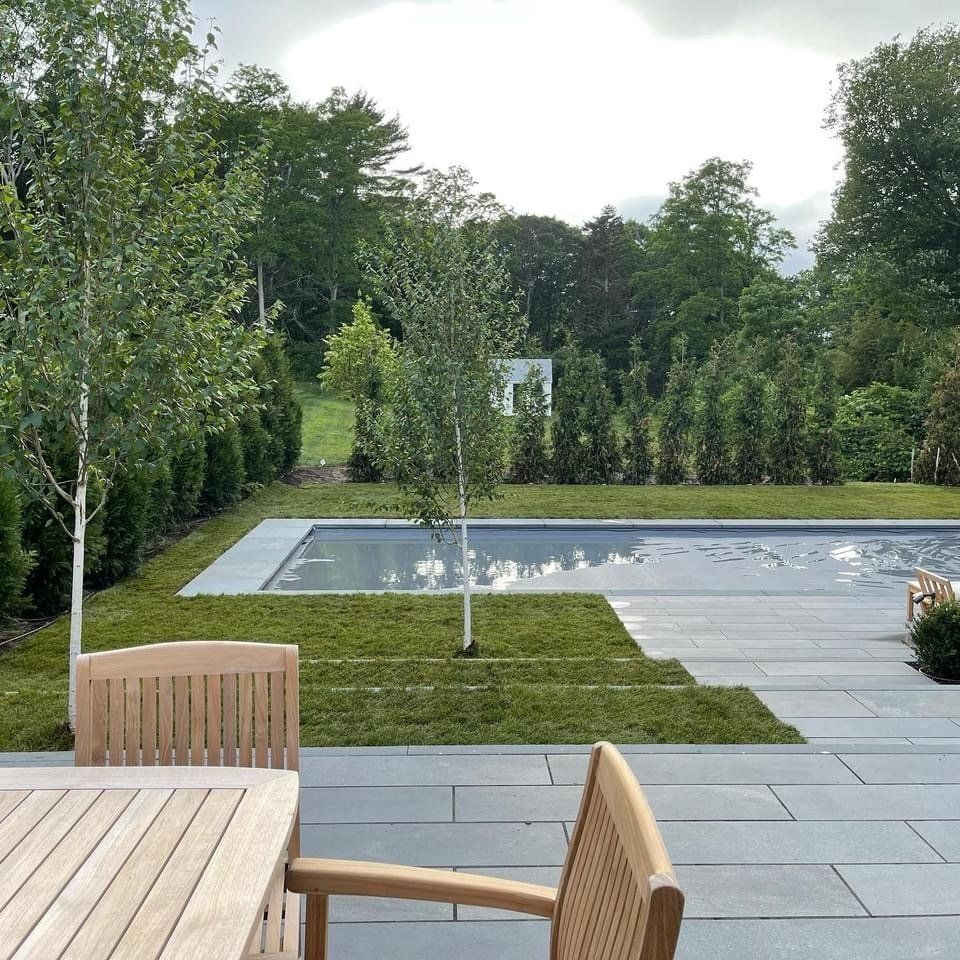 A teak patio set overlooks a backyard with a rectangular swimming pool, manicured lawn, birch trees, and tall hedges.