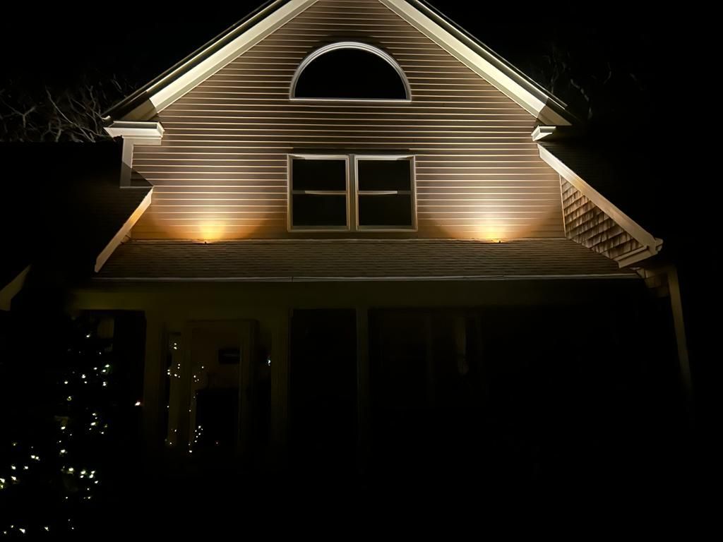A two-story house exterior at night with warm up-lighting highlighting the siding, gables, and a window.