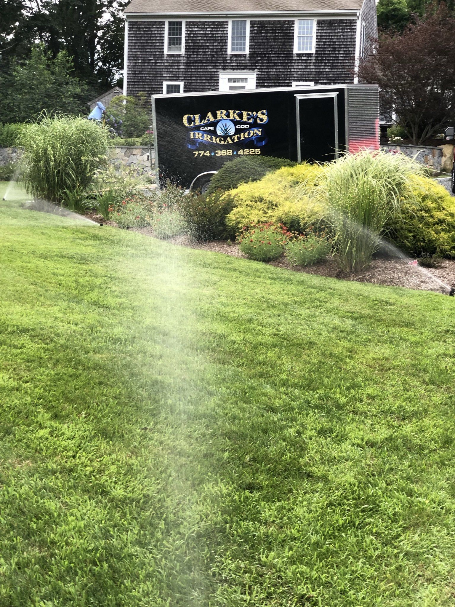 A lawn sprinkler sprays water over a green lawn in front of a dark trailer parked by a historic-looking stone house.