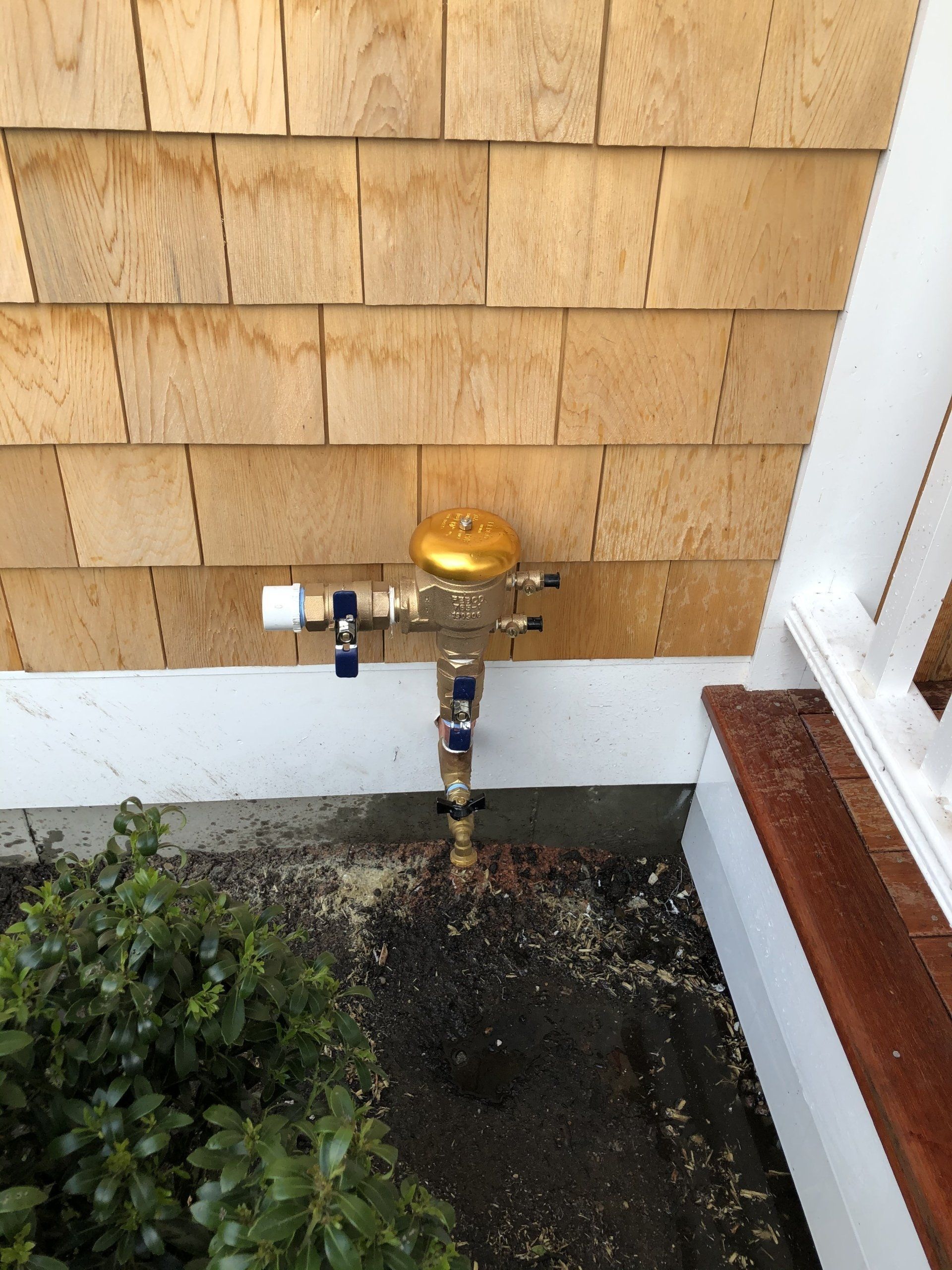 A backflow preventer assembly attached to the exterior shingled wall of a house near some plants and a wooden porch.
