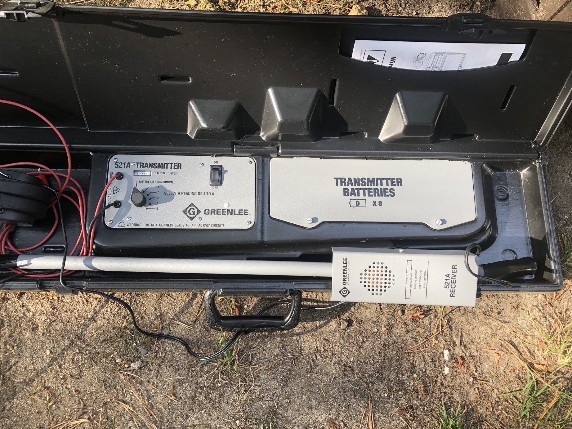 An open black case holding a Greenlee underground cable locator transmitter unit with battery compartment and leads.