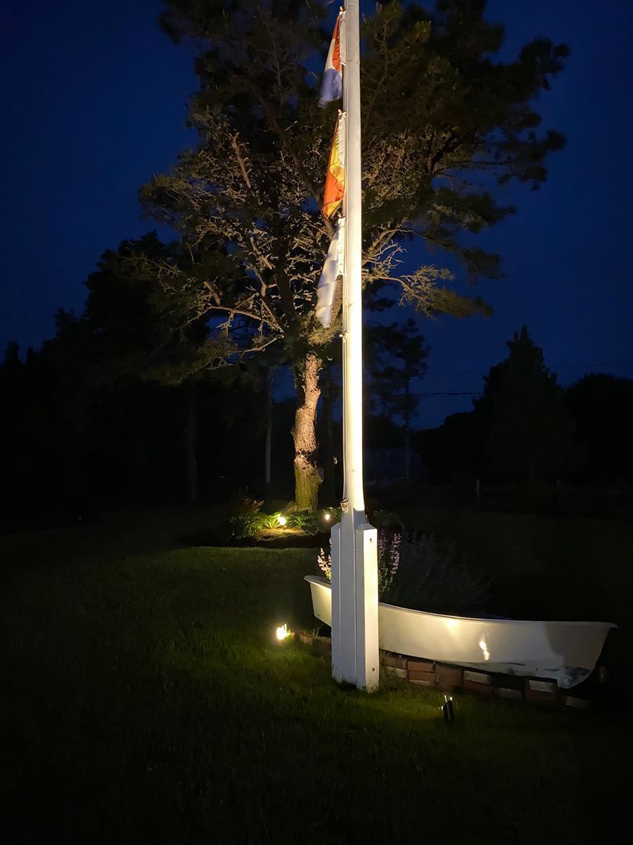 A white flagpole with multiple flags, illuminated at night, stands near a small boat in a grassy area with trees behind.