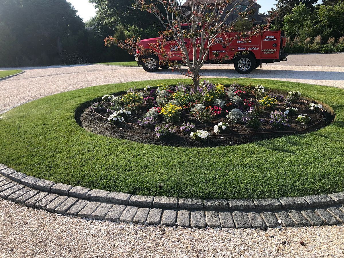 A circular garden bed with flowers and a small tree, bordered by cobblestones and surrounded by a white gravel driveway.
