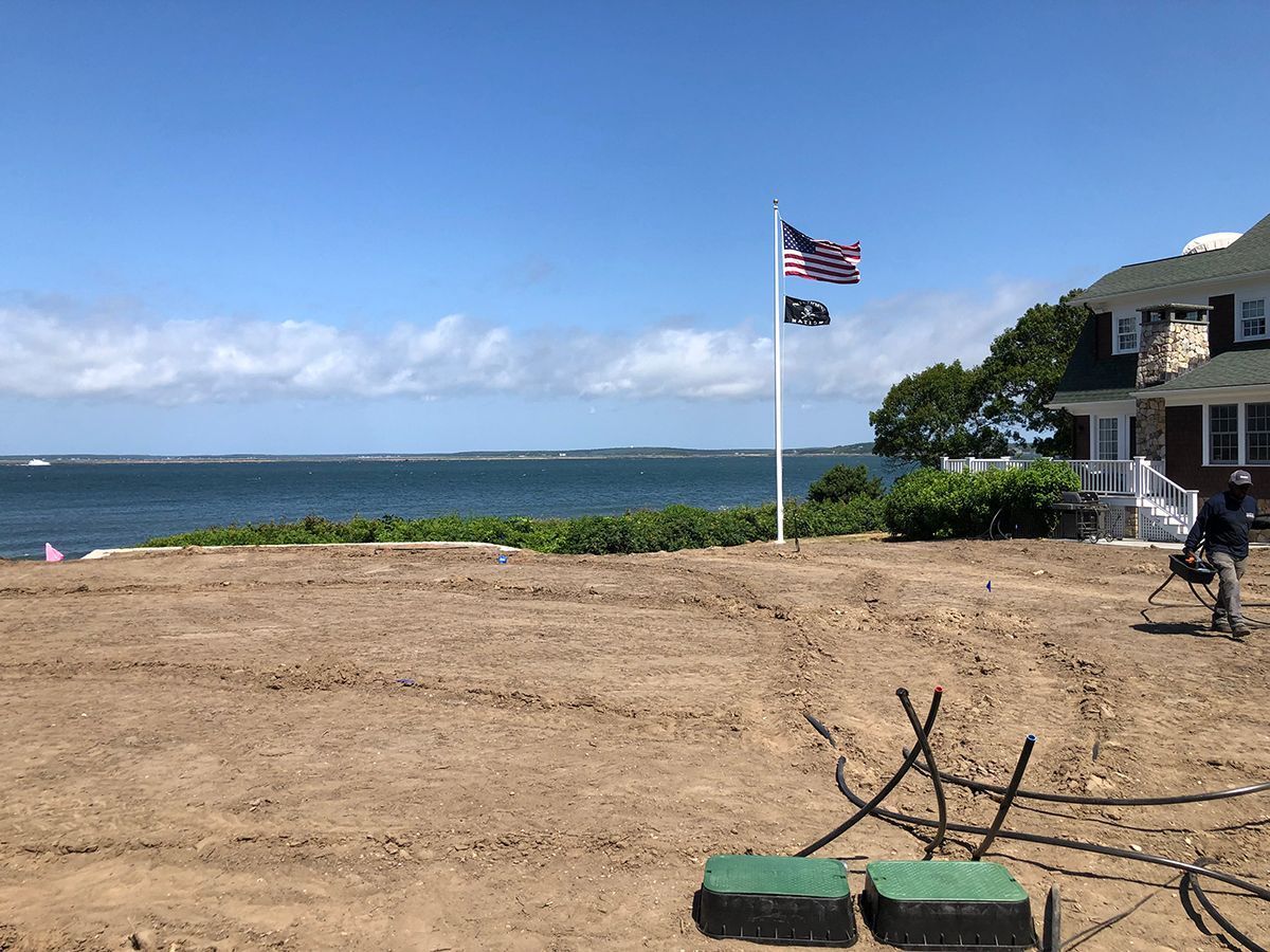 A person stands on a dirt construction site overlooking a bay with a flag pole and house nearby under a clear blue sky.