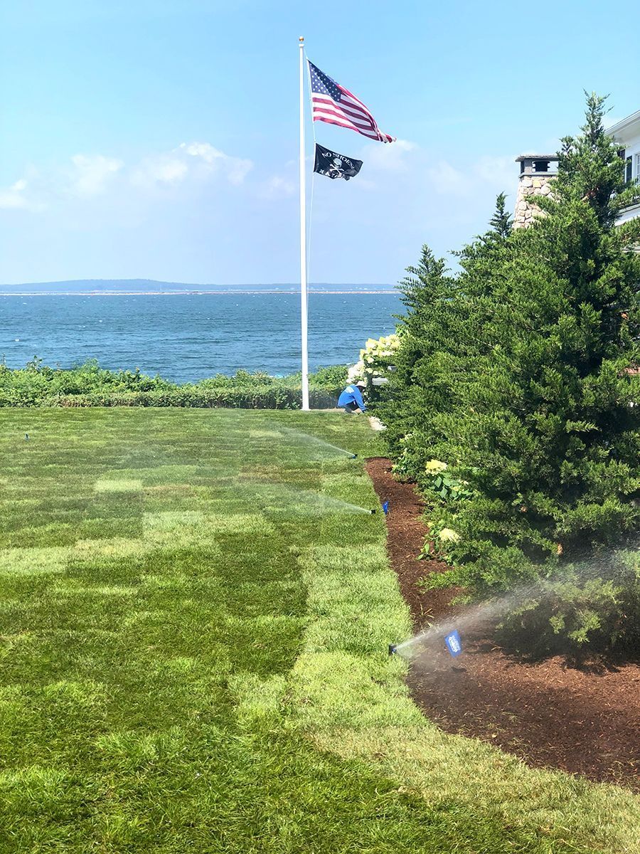 A flag pole with the American and POW/MIA flags stands on a grassy lawn beside a seaside house with sprinklers running.