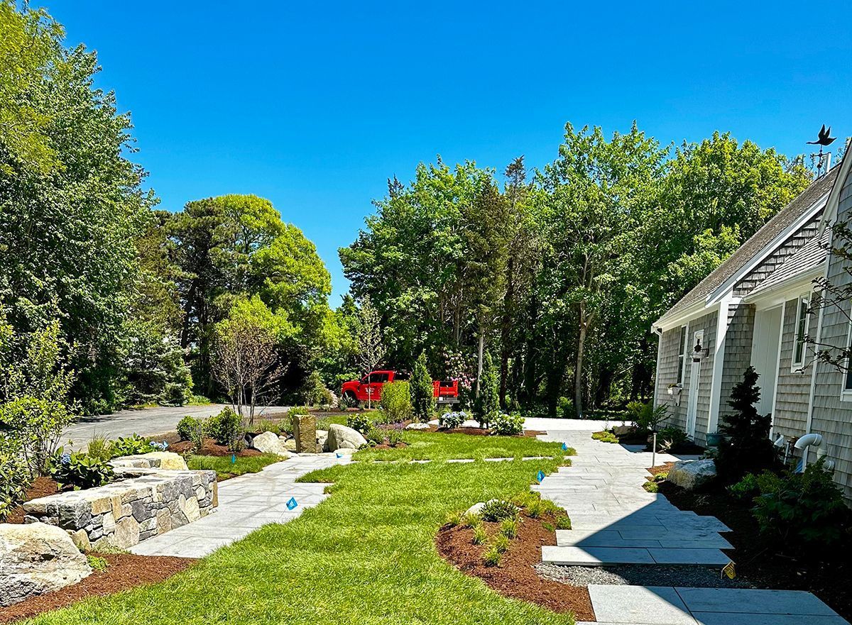 A stone pathway winds through a grassy lawn between a stone cottage and a retaining wall, bordered by trees under a blue sky.
