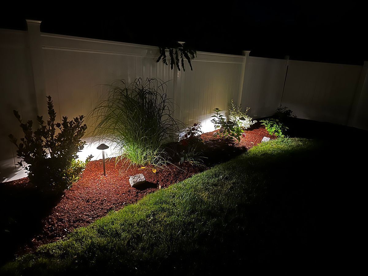 A manicured backyard garden bed at night, illuminated by small landscape lights against a white vinyl privacy fence.