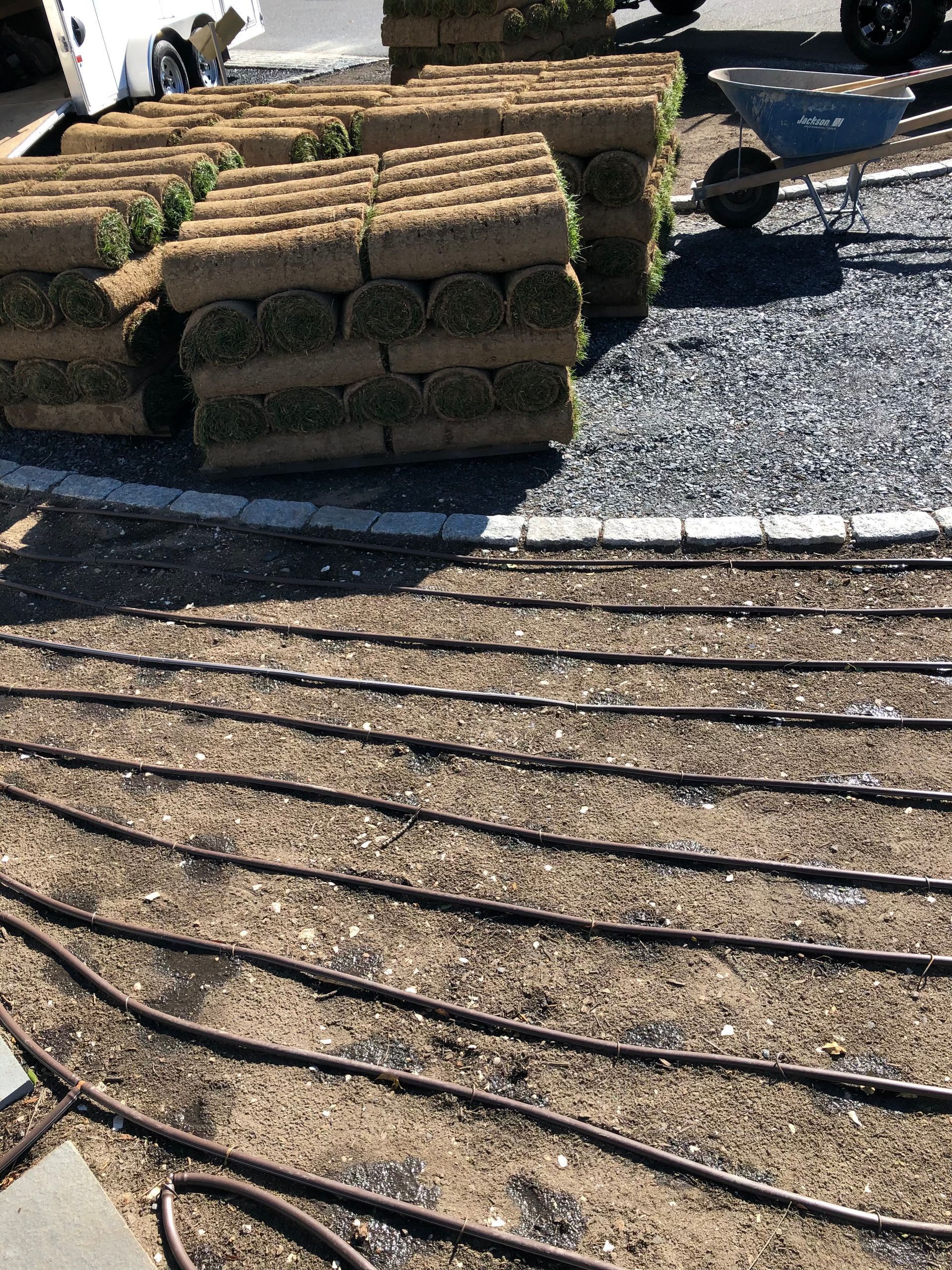 Stacked pallets of sod sit on gravel beside a yard prepared with black irrigation tubing for a landscaping project.