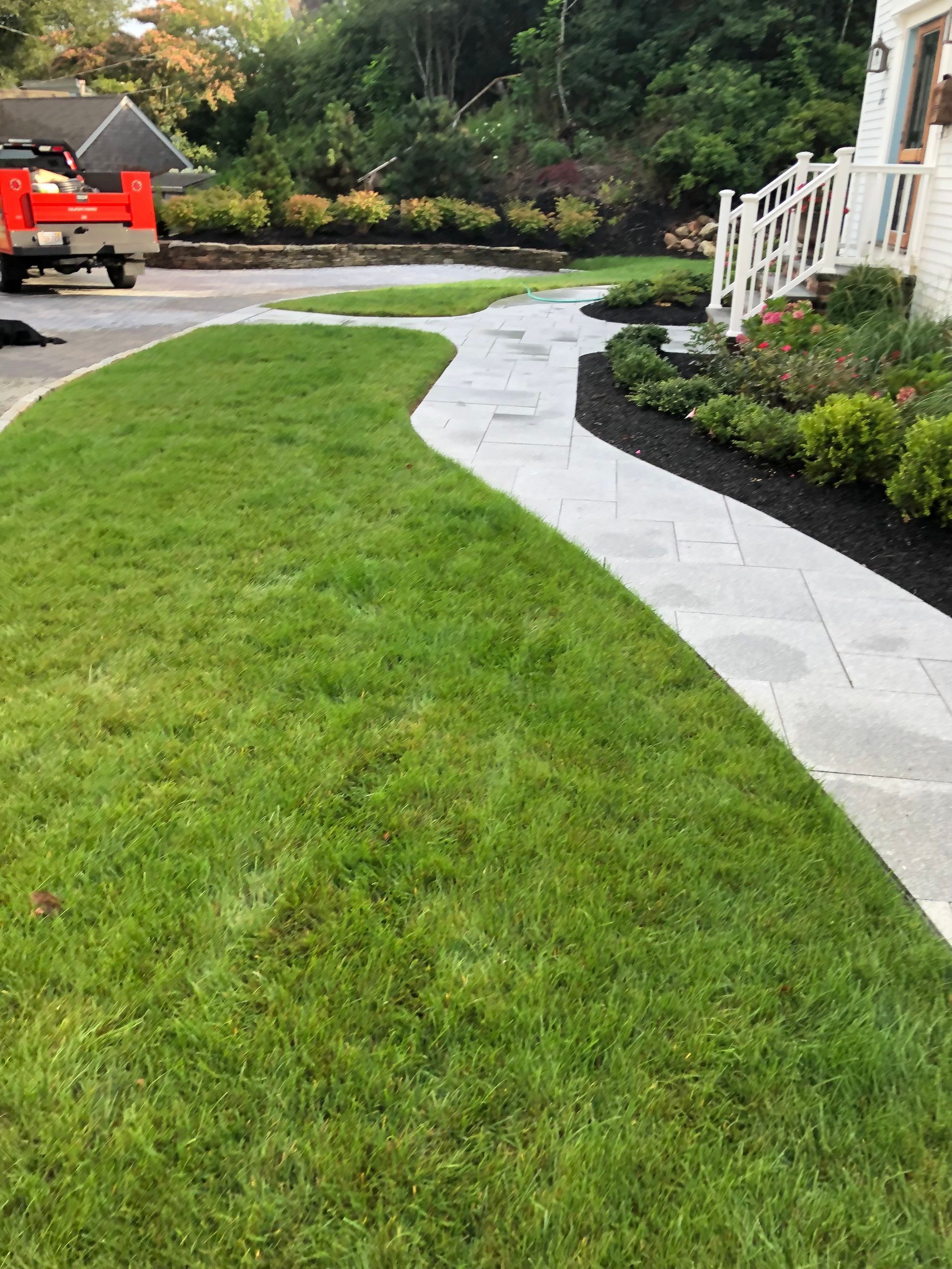 A curved light grey stone walkway leads to a house porch, framed by a green lawn and dark mulch garden beds.