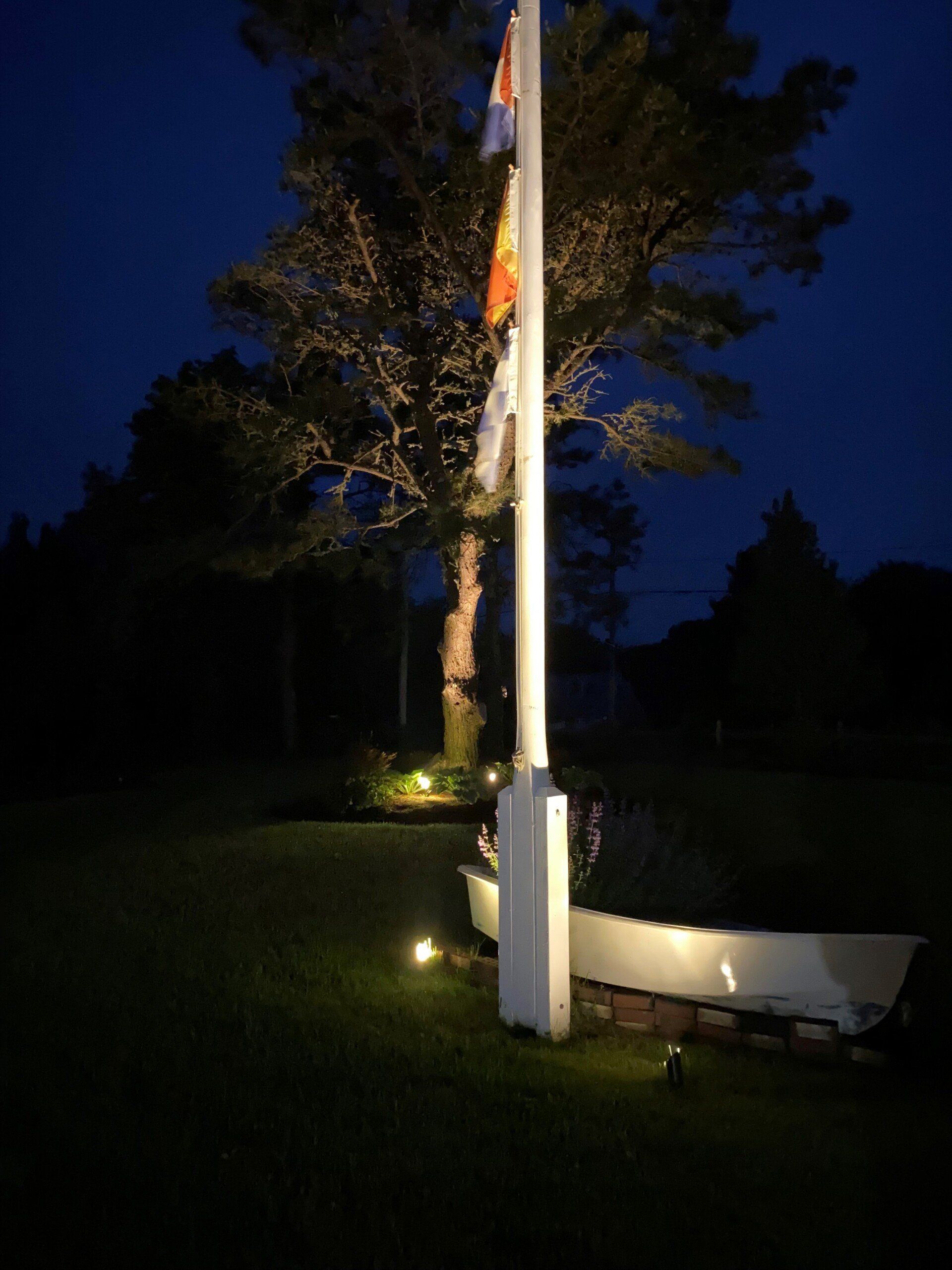 A flagpole with multiple flags stands illuminated at night in a garden with a small boat planter at its base.