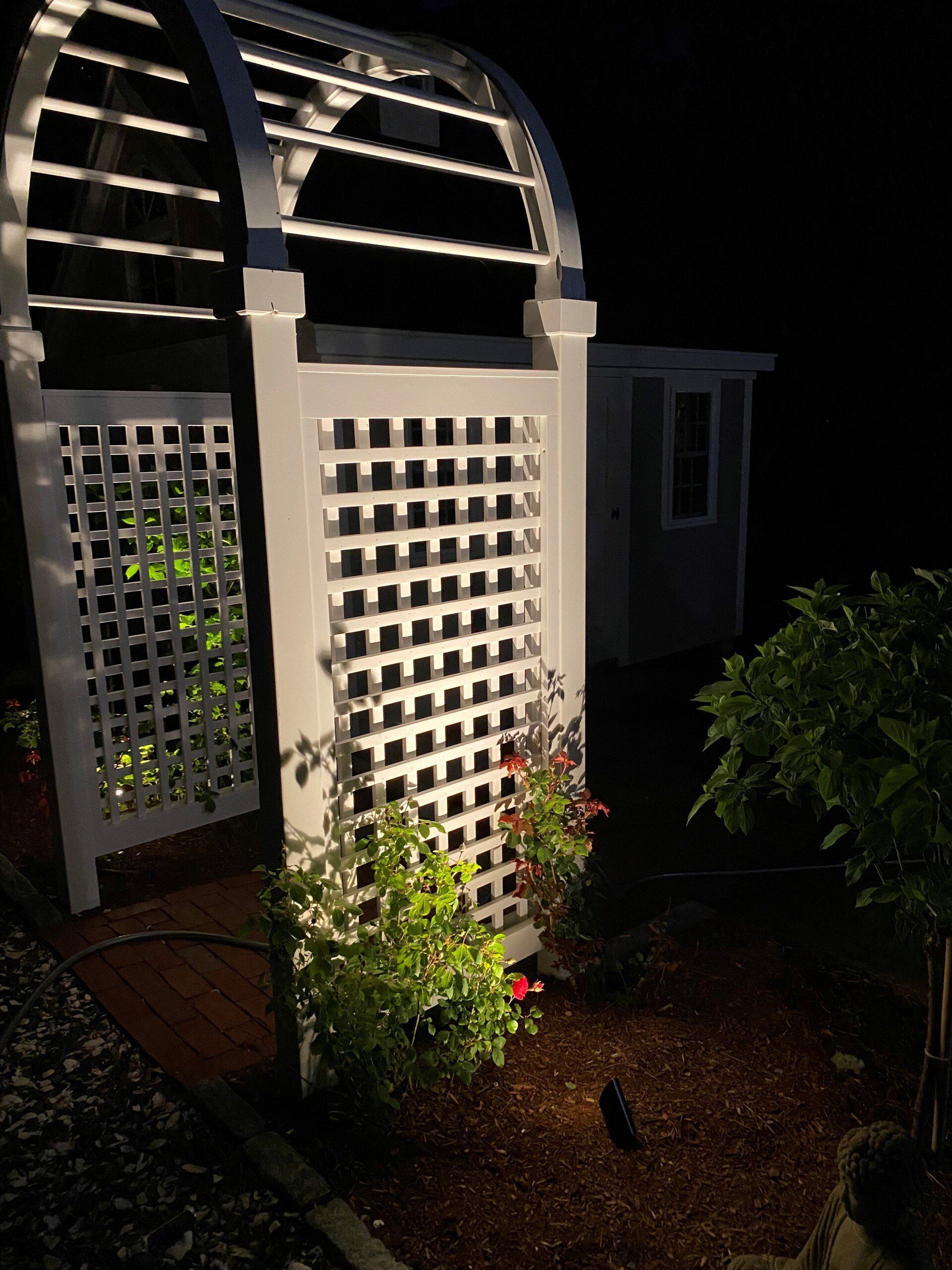 A white wooden garden arbor illuminated at night, surrounded by mulch, greenery, and a shed in the background.