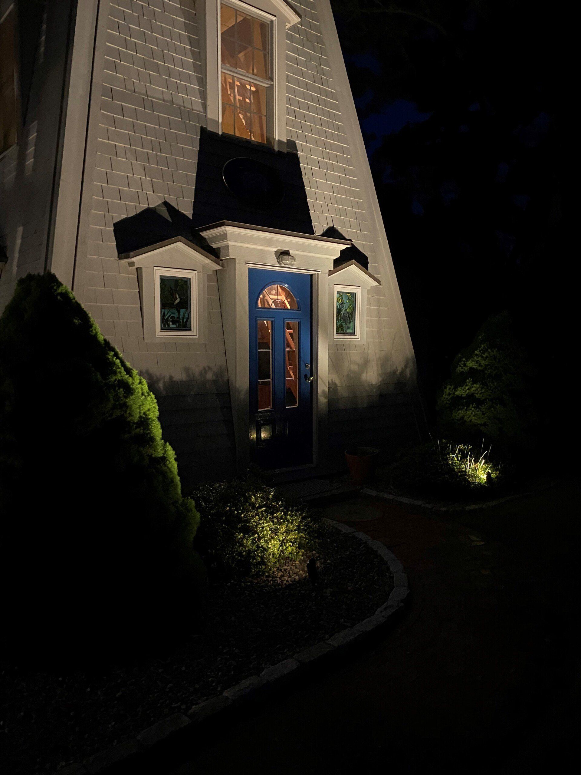 An A-frame house entrance at night with a blue door, lit by exterior spotlights and landscaping lights.