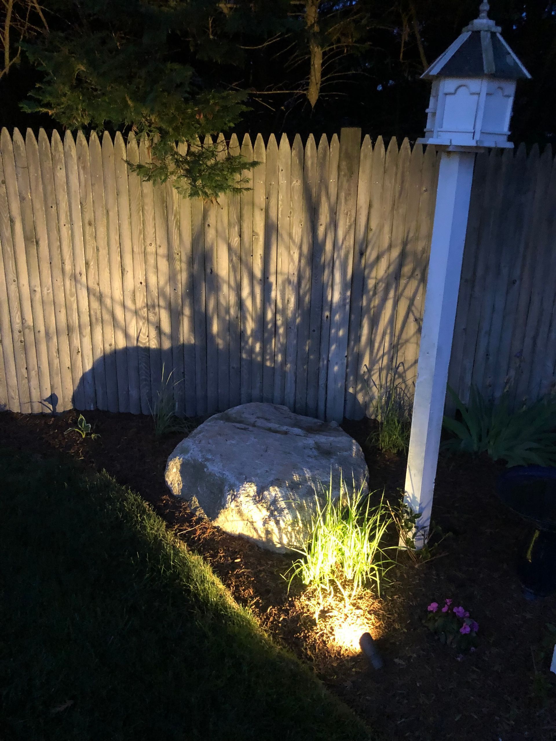 A large rock and birdhouse illuminated by a ground light at night in front of a wooden fence.