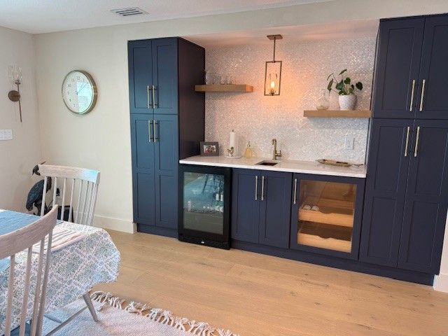 Blue cabinets in a kitchen area with a small sink, wine cooler, and decorative tile backsplash.