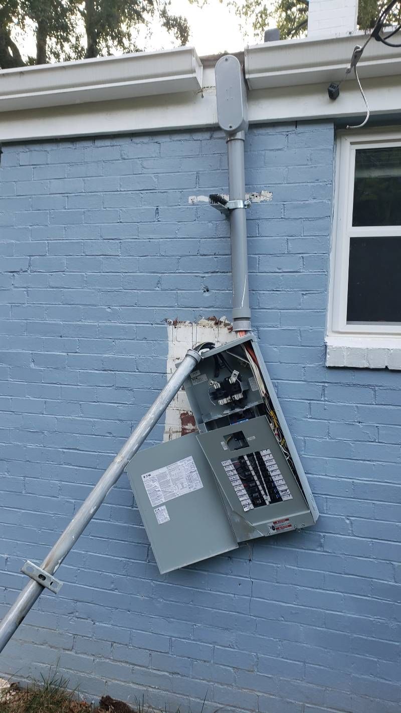 A broken electrical box is sitting on the side of a blue brick house.