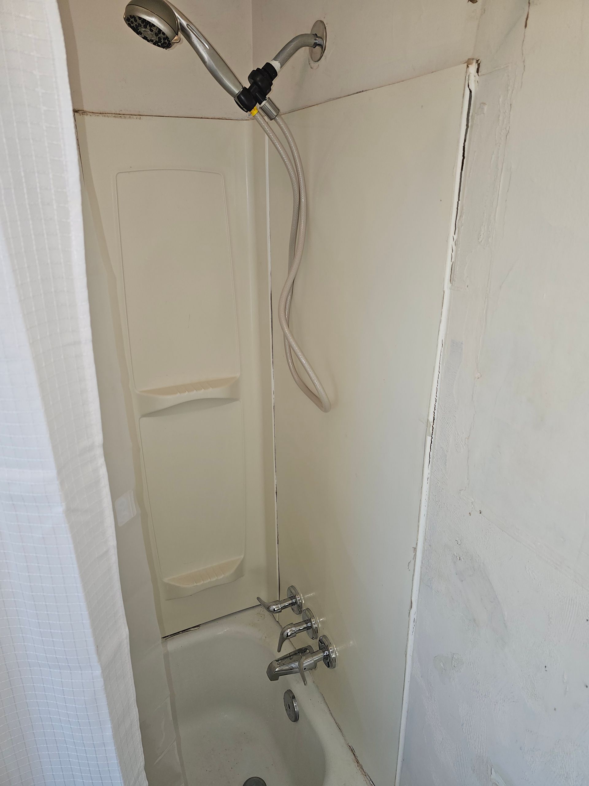 A small, off-white shower stall with a handheld showerhead and a bathtub.
