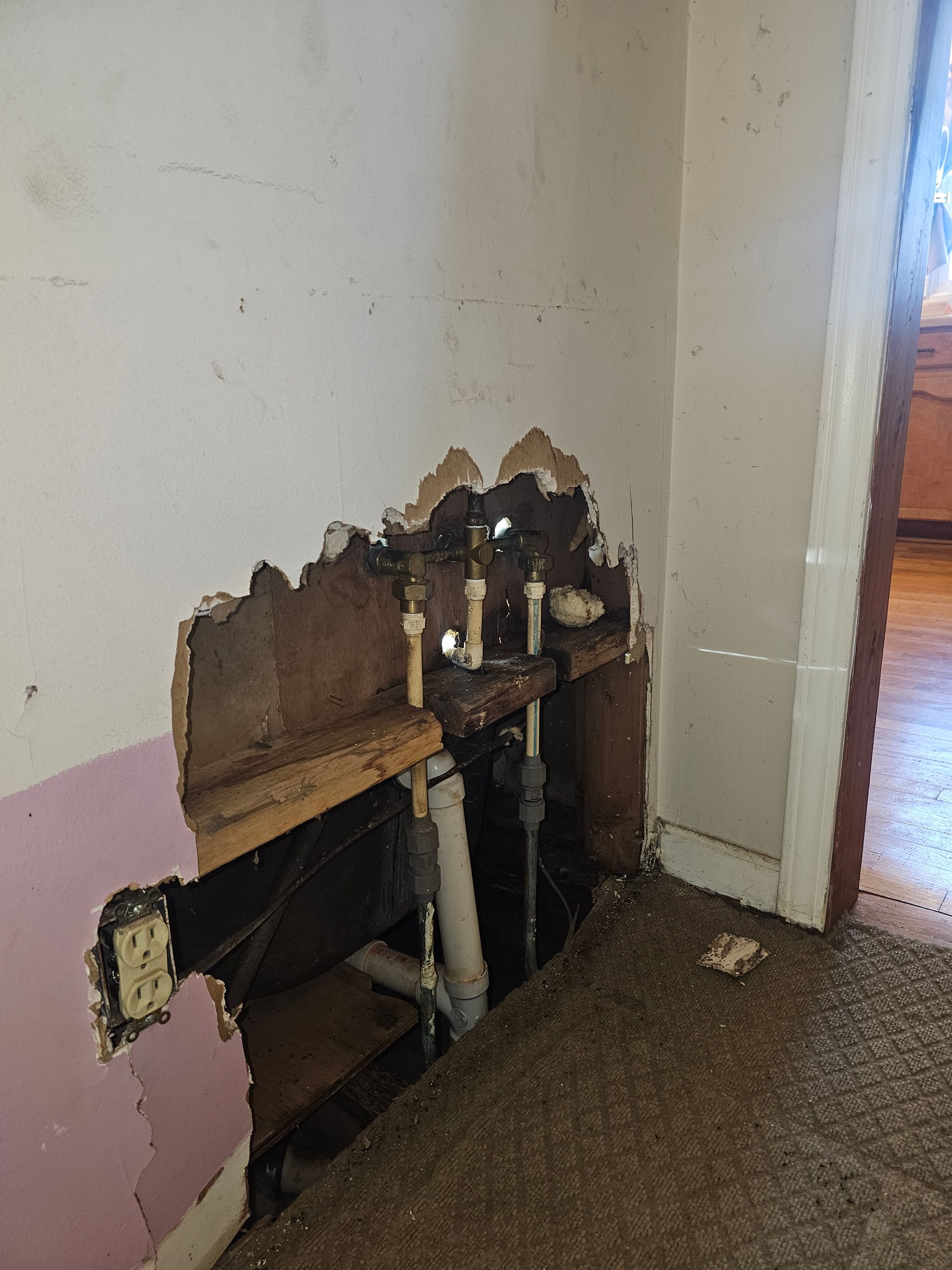 Hole in wall reveals plumbing pipes and electrical outlet; damaged wall with brown rot and pink paint.