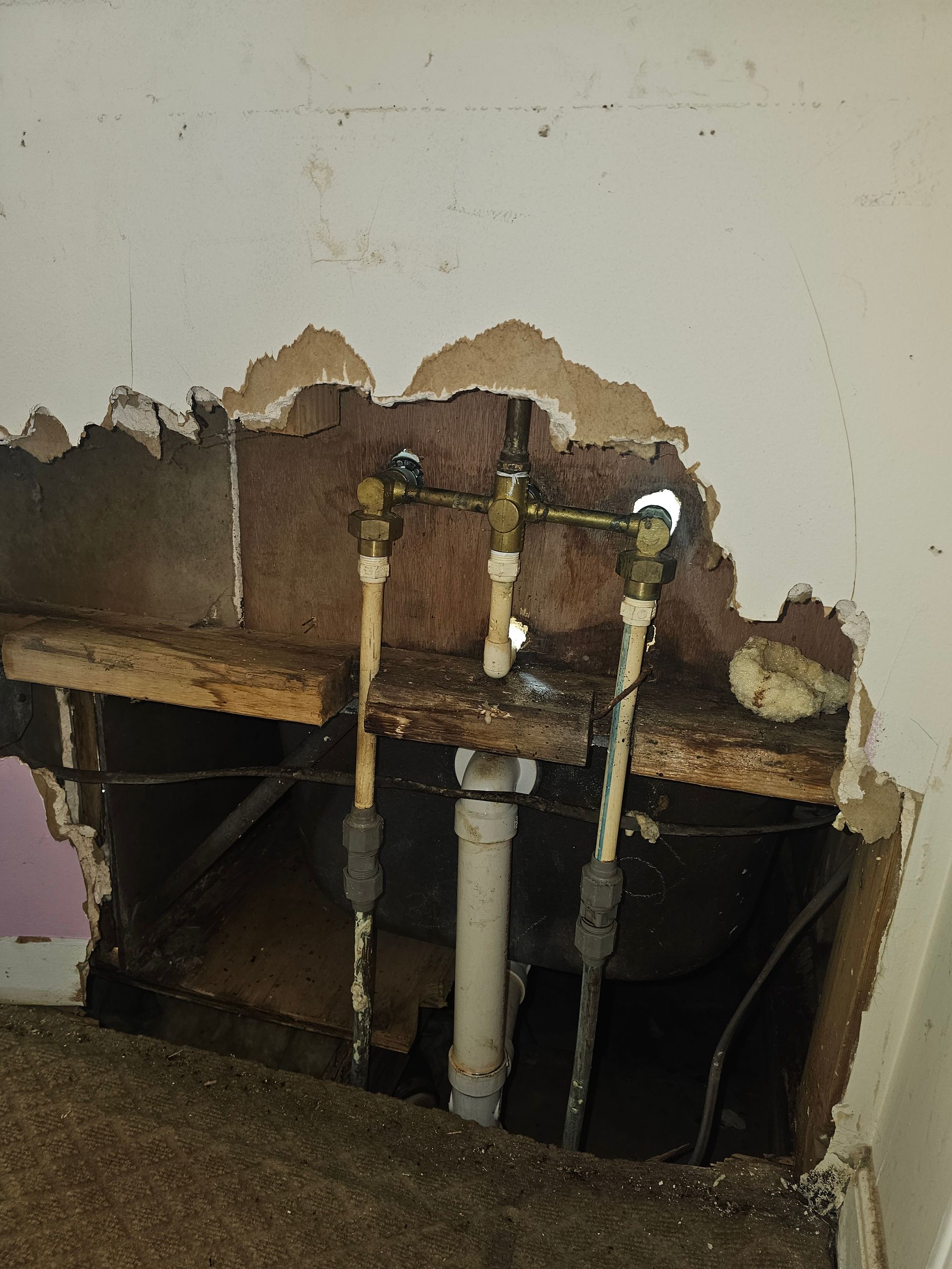 Pipes exposed in a hole in a wall; water valves and drain visible. Brown wood and damaged drywall surround.