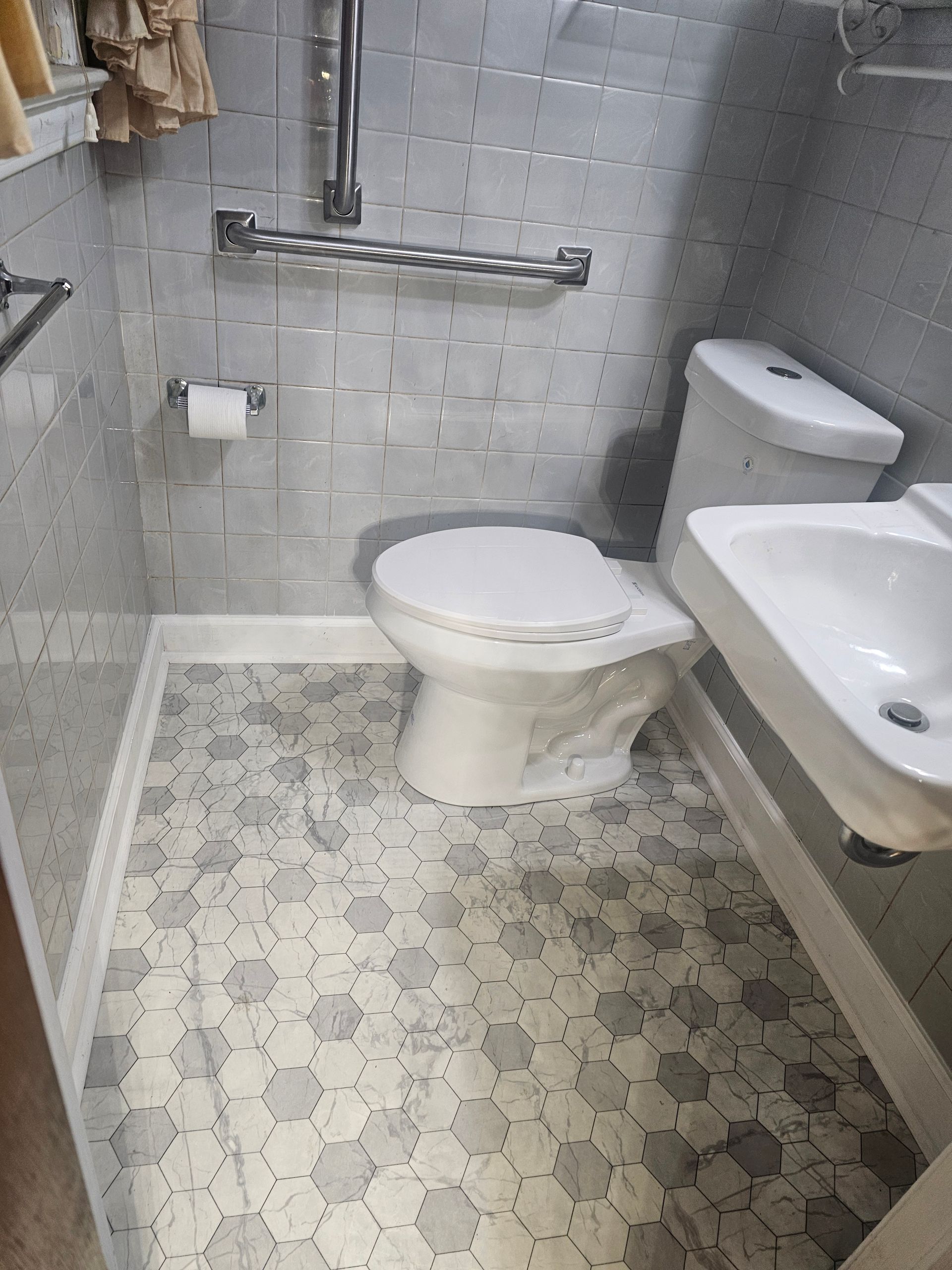 Small bathroom with toilet, sink, and safety grab bars; hexagon tile floor and white-gray tiled walls.