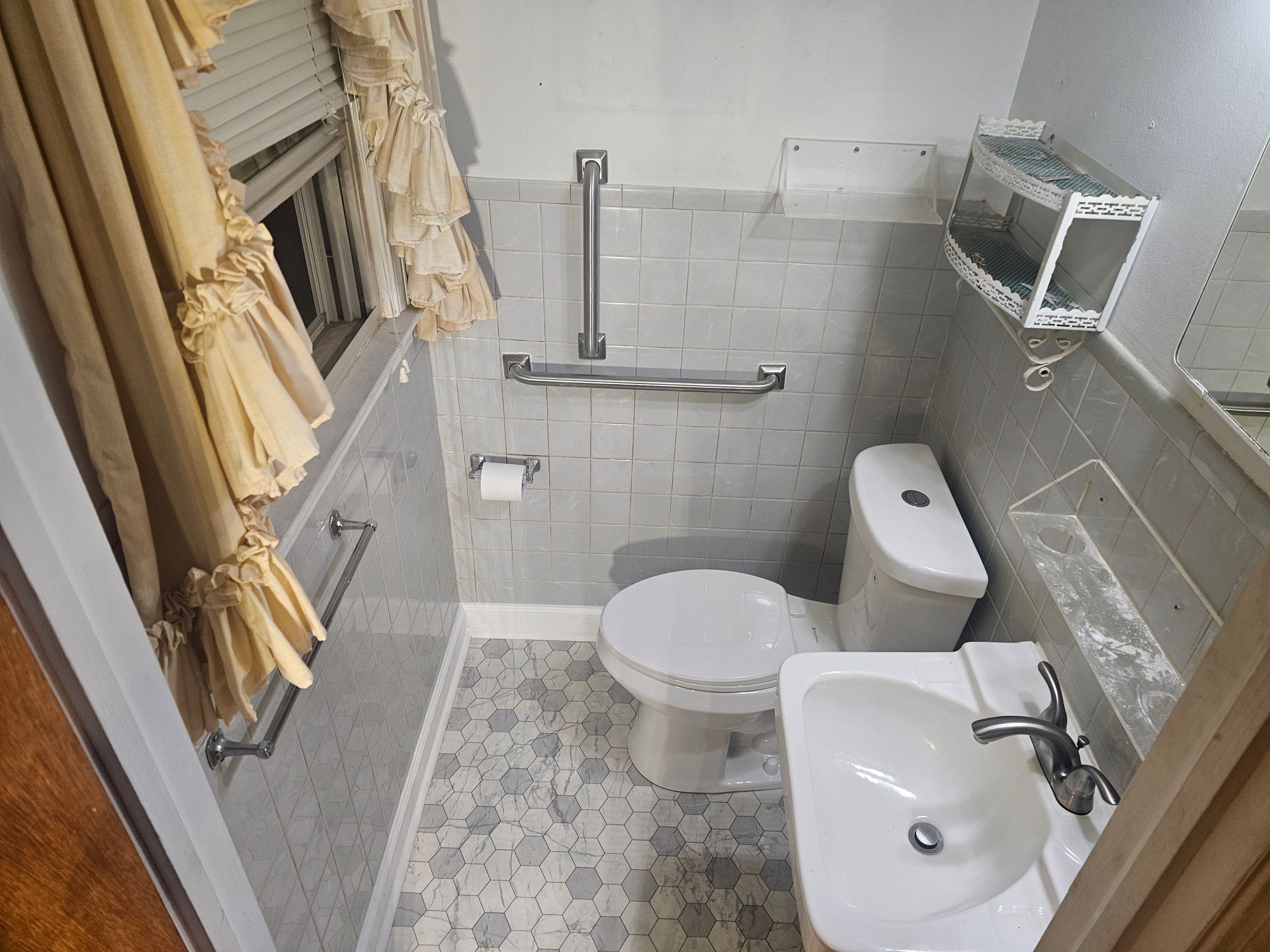 Small, tiled bathroom with toilet, sink, grab bars, and floral-patterned floor. White and gray tones.