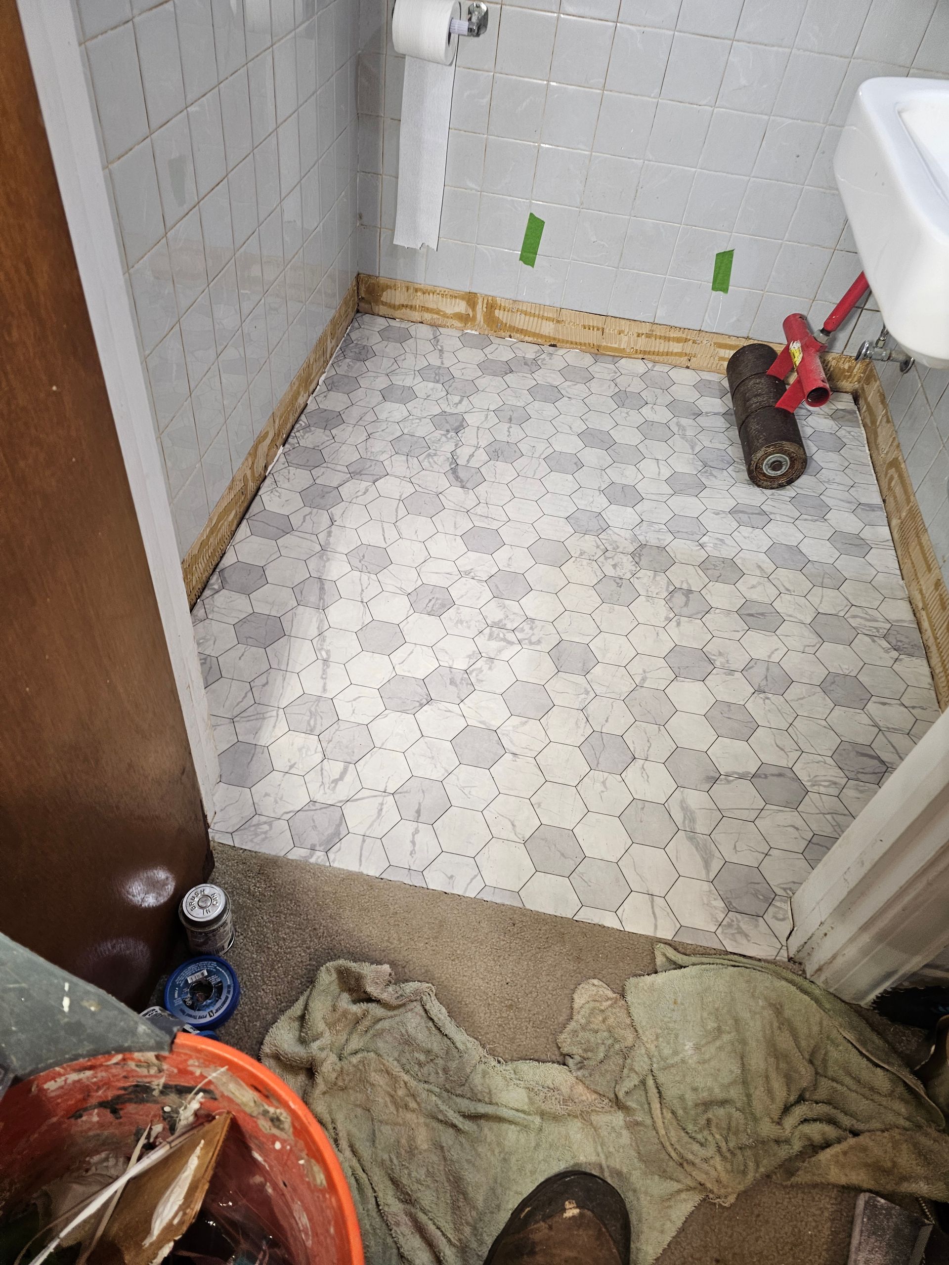 Bathroom floor with patterned vinyl being installed. Roller, wood trim, and supplies present.