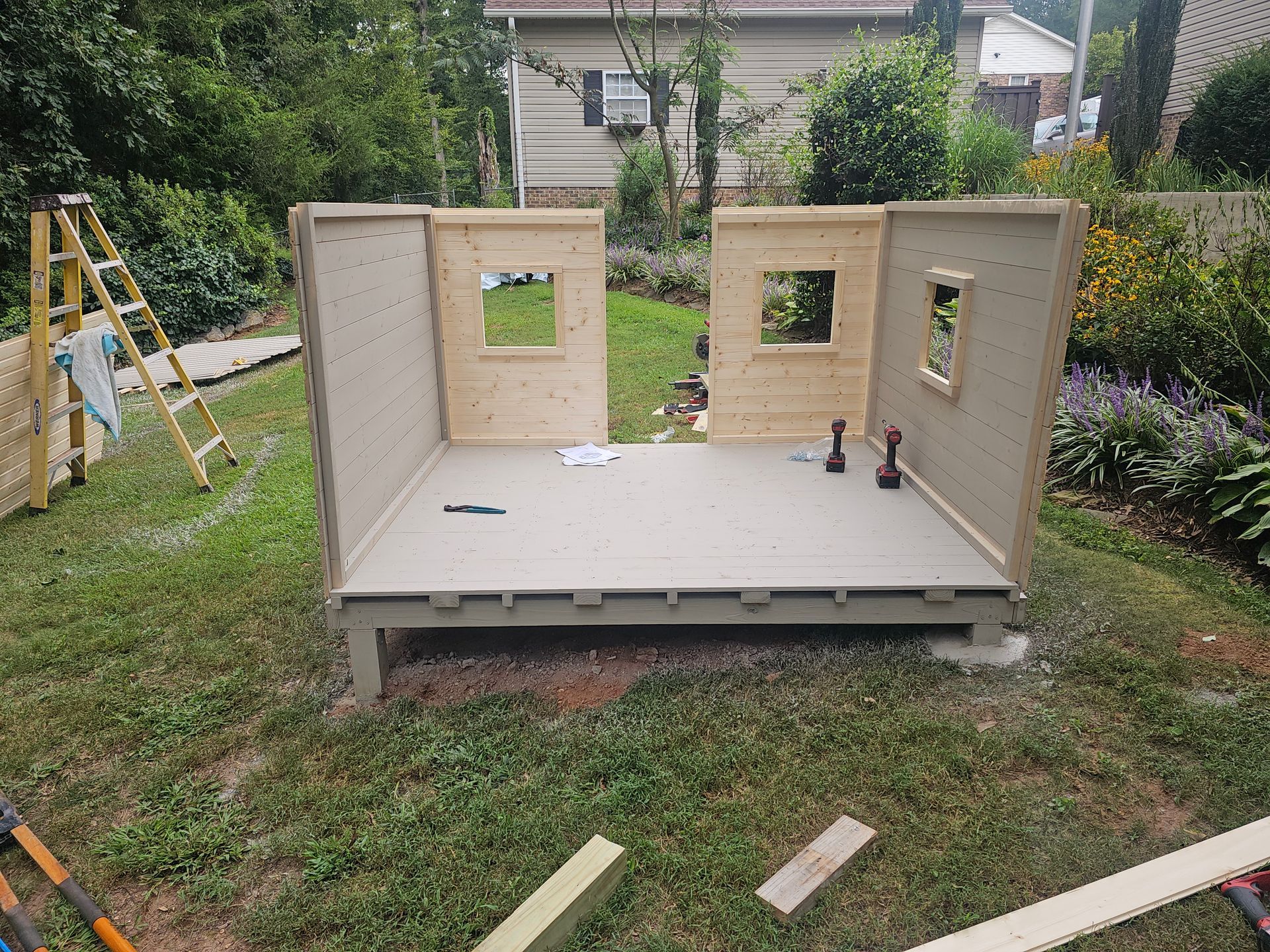 Partially built small wooden shed with window cutouts on a grassy yard, boards and a ladder nearby