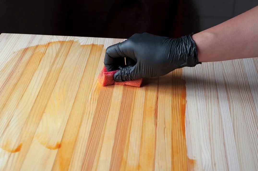 A person wearing black gloves is cleaning a wooden surface with a sponge.