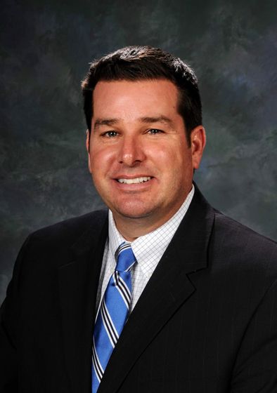 Portrait of a smiling man in a black suit and blue striped tie against a studio backdrop