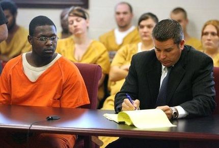 Defendant in orange jail uniform sits beside attorney taking notes in a courtroom, with spectators behind them.