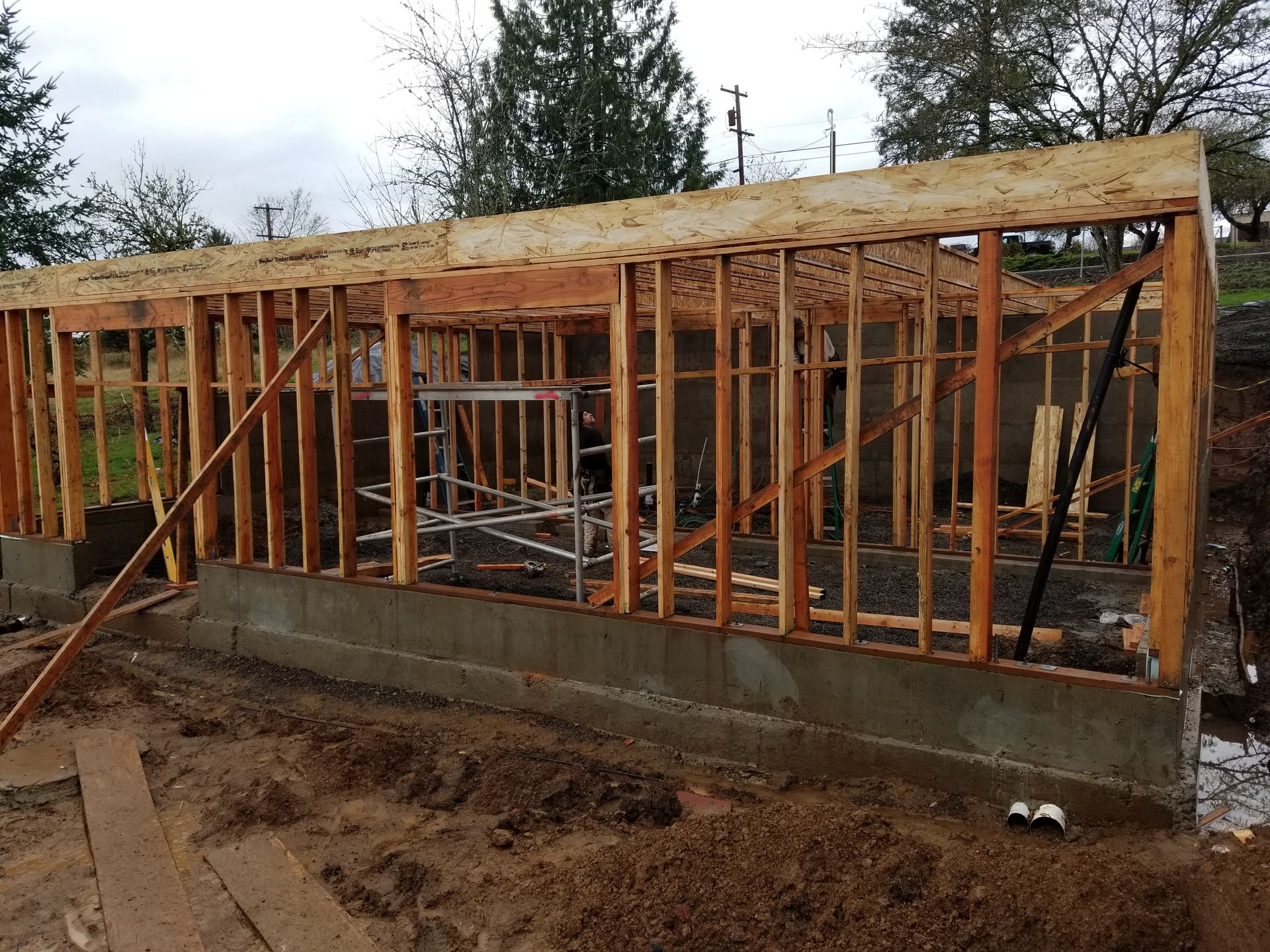 Wood-framed house under construction on a concrete foundation, with muddy ground in front.