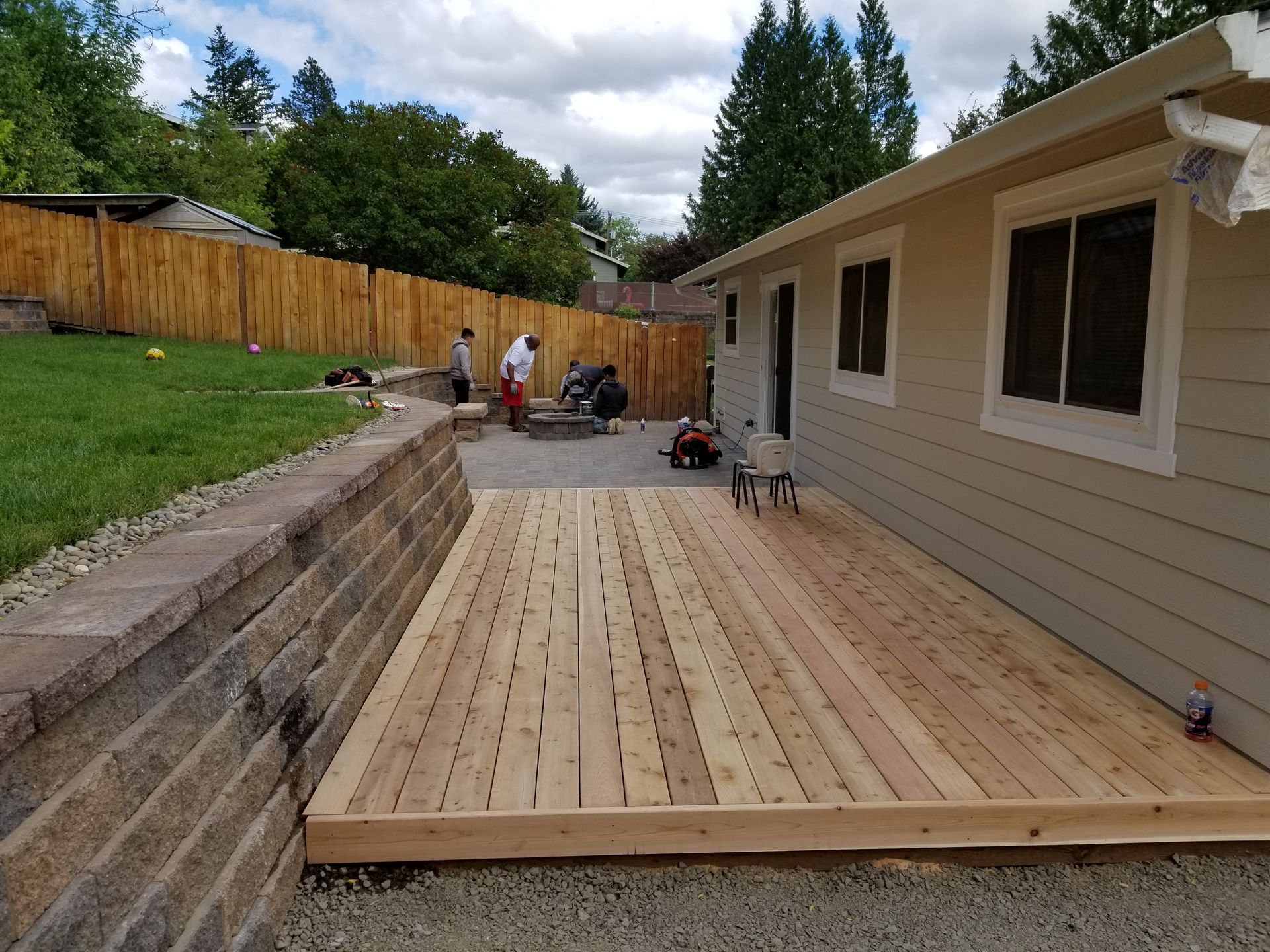 Backyard wooden deck under construction beside a house, with workers and tools near a fenced yard