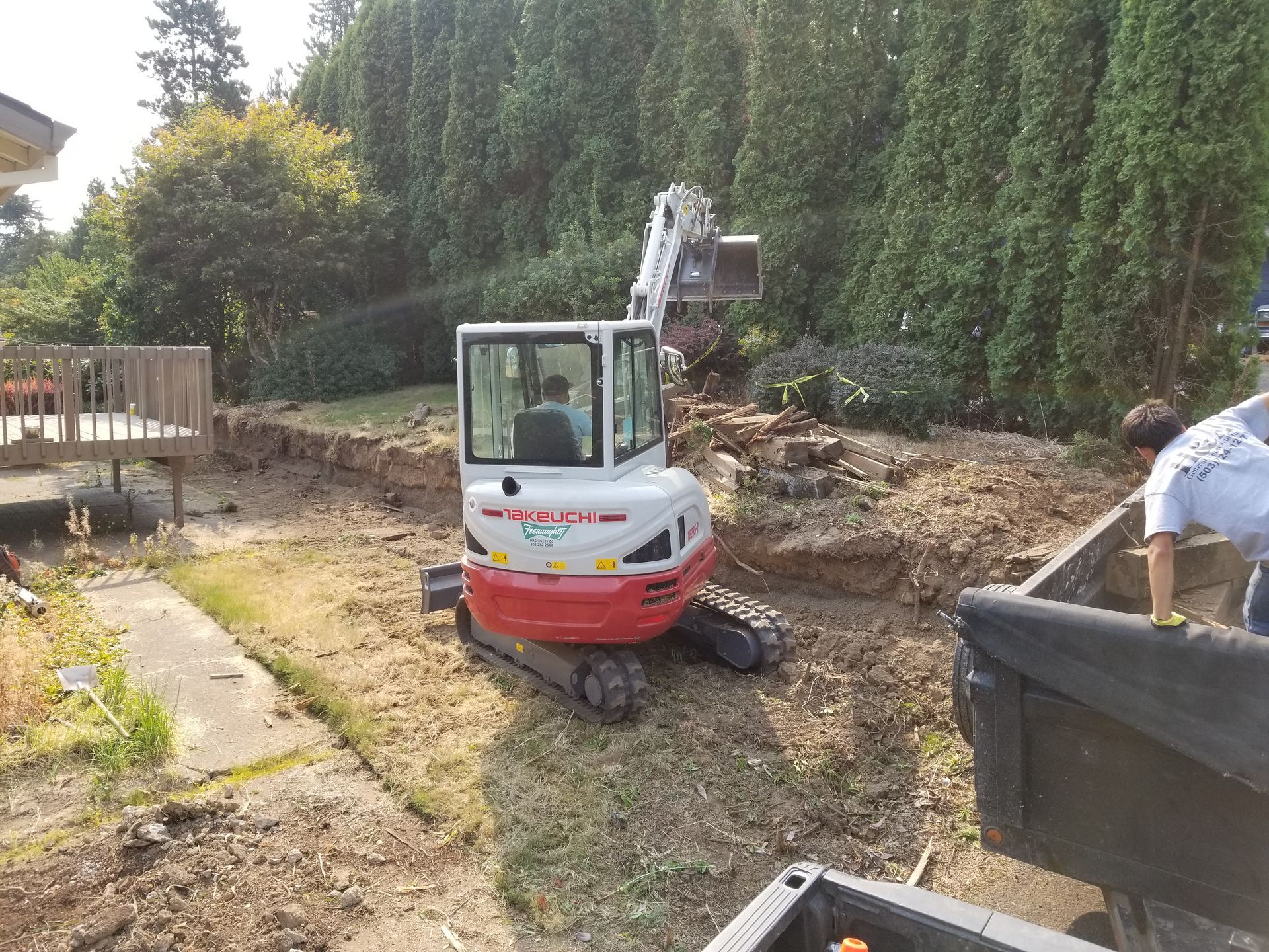 Mini excavator working in a backyard excavation area, with a person digging nearby.