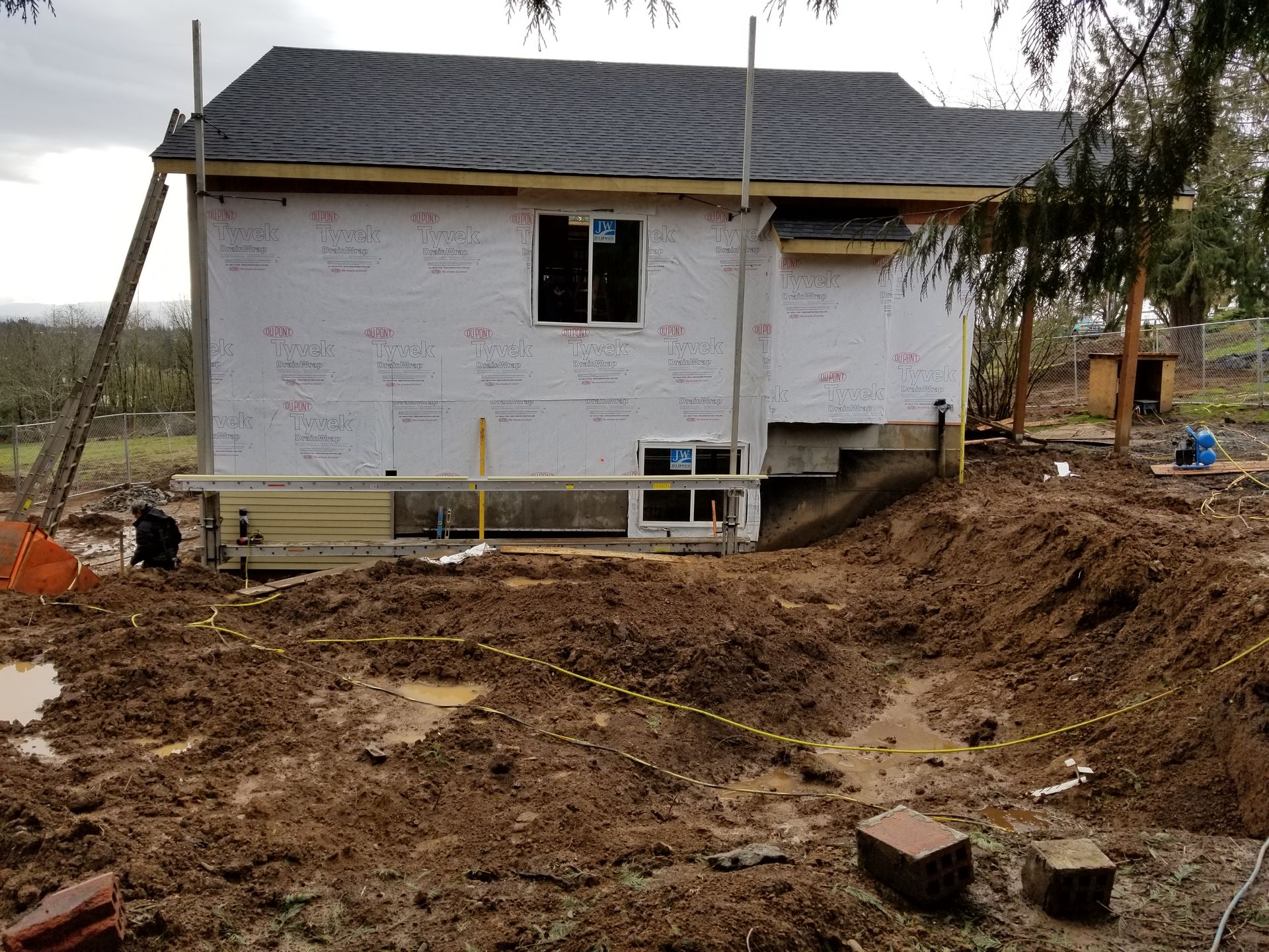Small house under construction on a muddy site, with white wrap, exposed framing, and scattered bricks.