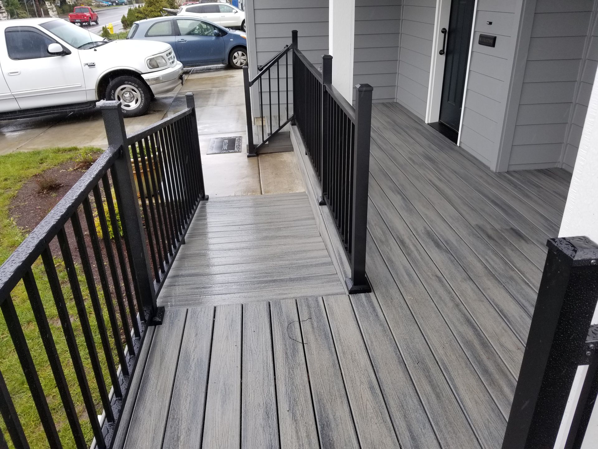 Black wooden ramp with handrails leading to a house entrance on a rainy day, with parked cars nearby