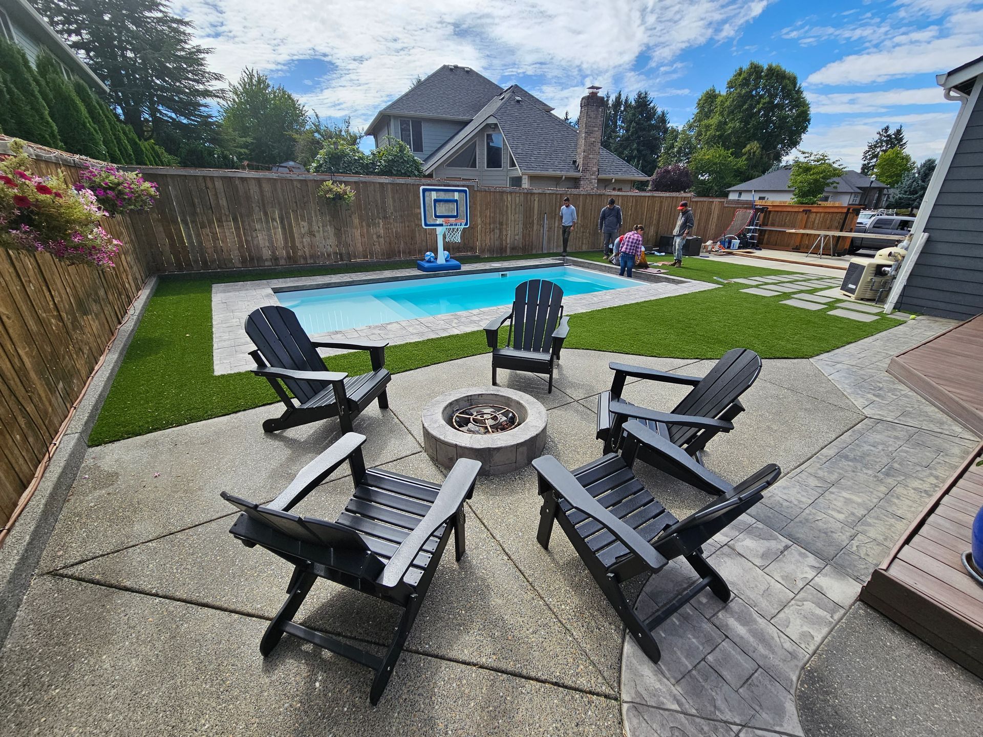 Backyard patio with fire pit chairs, lawn, pool, and basketball hoop under a blue sky.