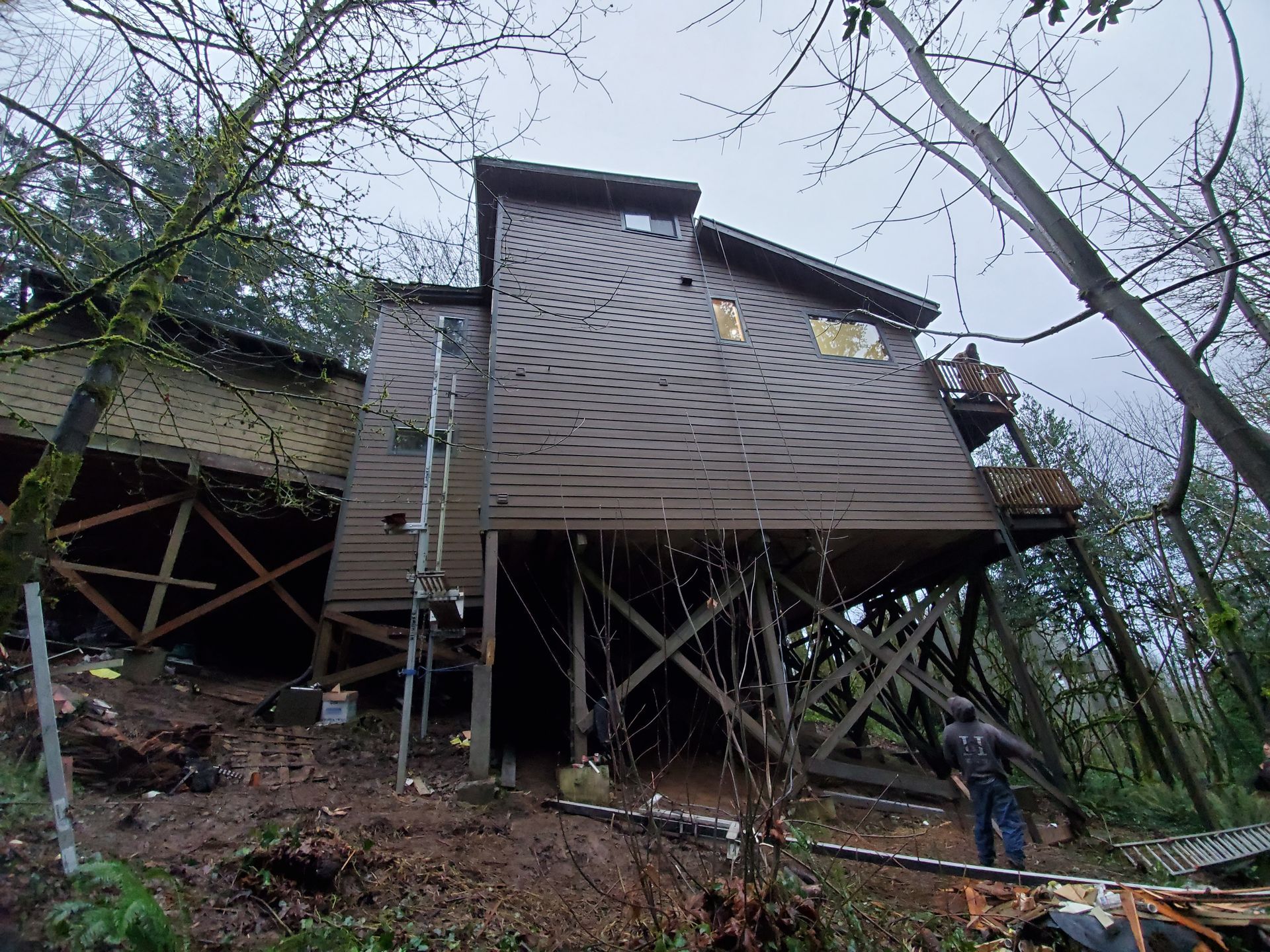 Elevated wooden house on stilts in a wooded hillside, with a ladder and cross-braced supports below.