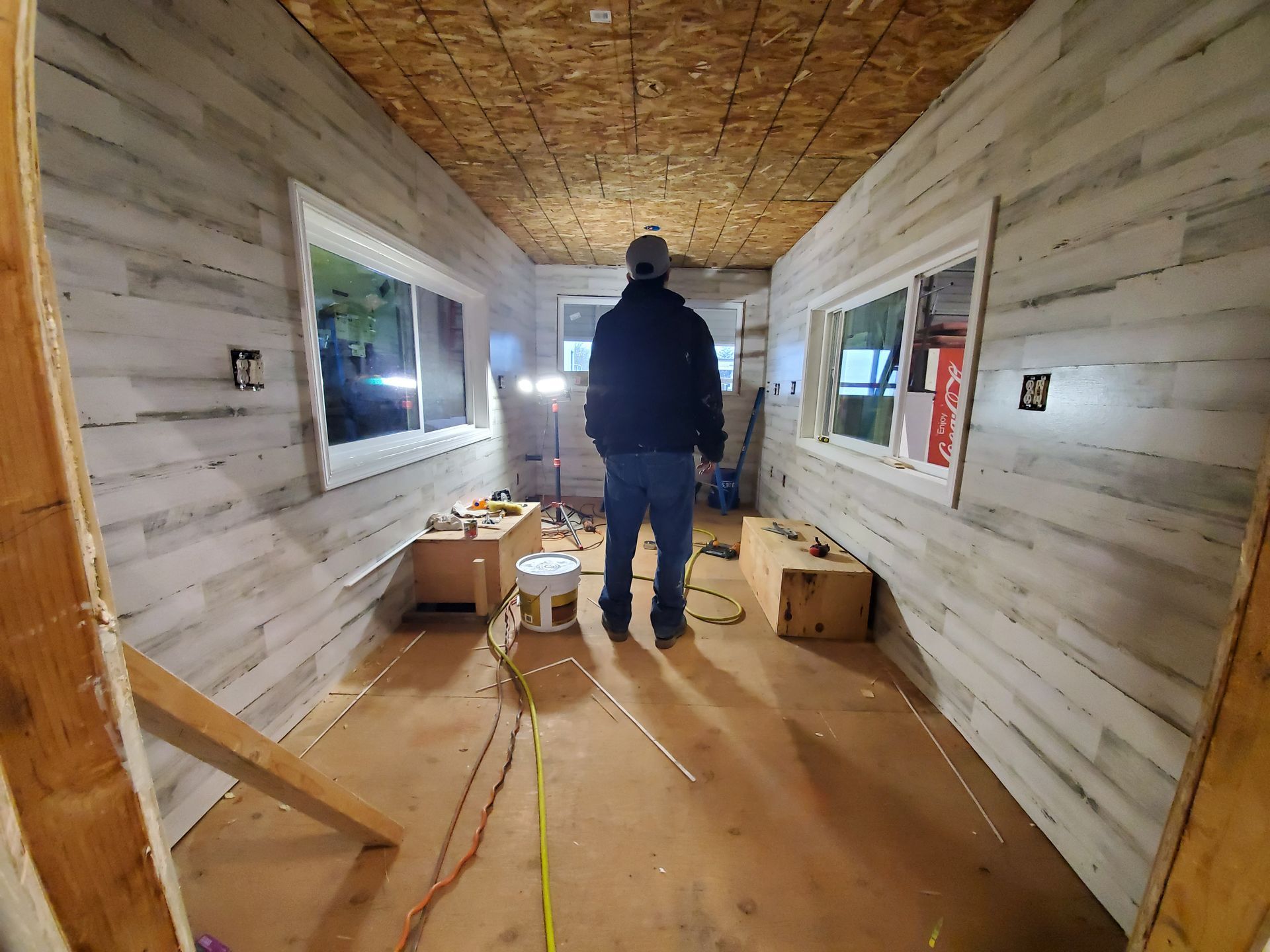 Person standing in a narrow, unfinished hallway with wood-paneled walls and windows, surrounded by construction materials.