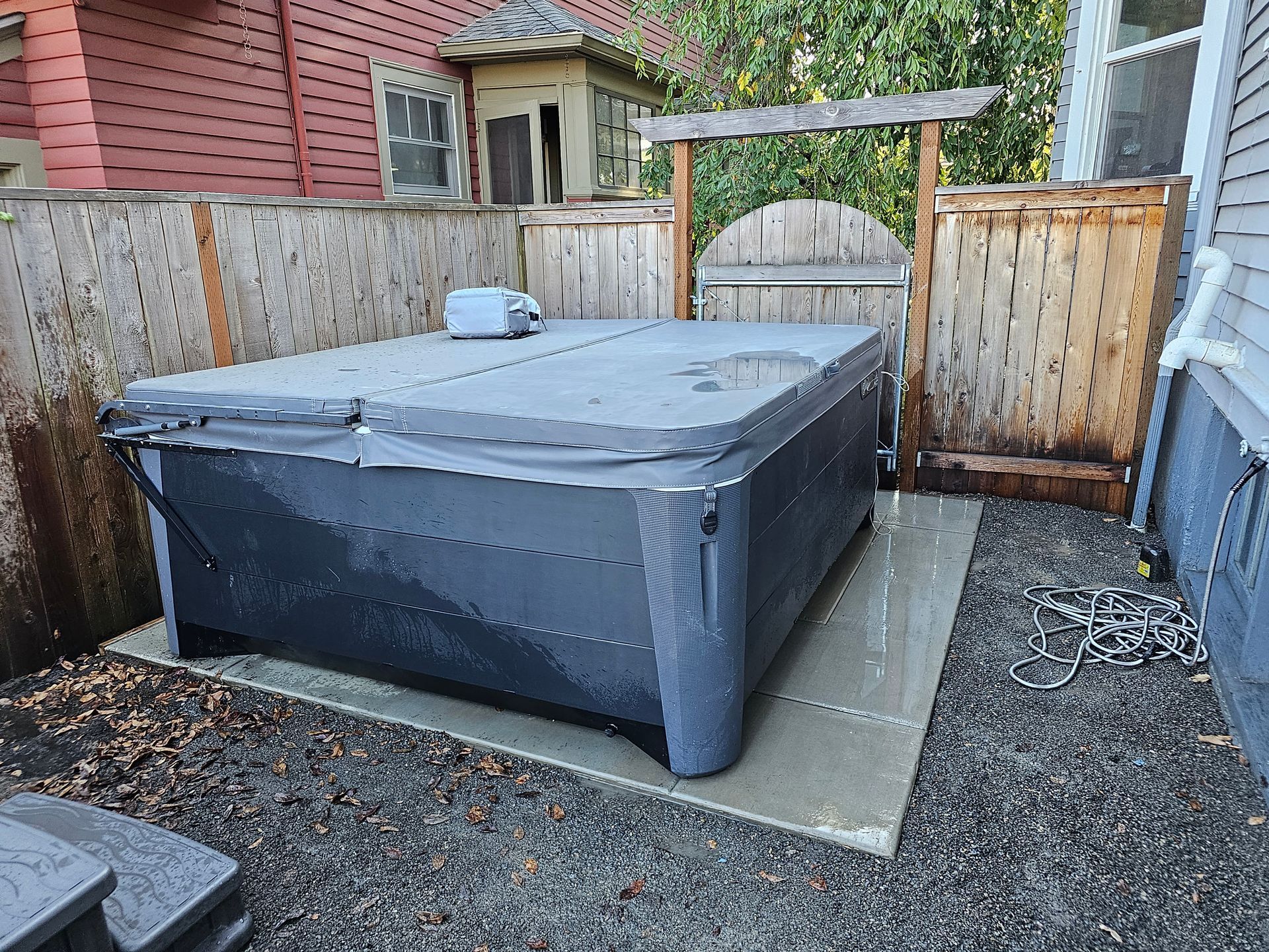 Covered hot tub in fenced backyard on a concrete pad, surrounded by gravel and wood fencing.
