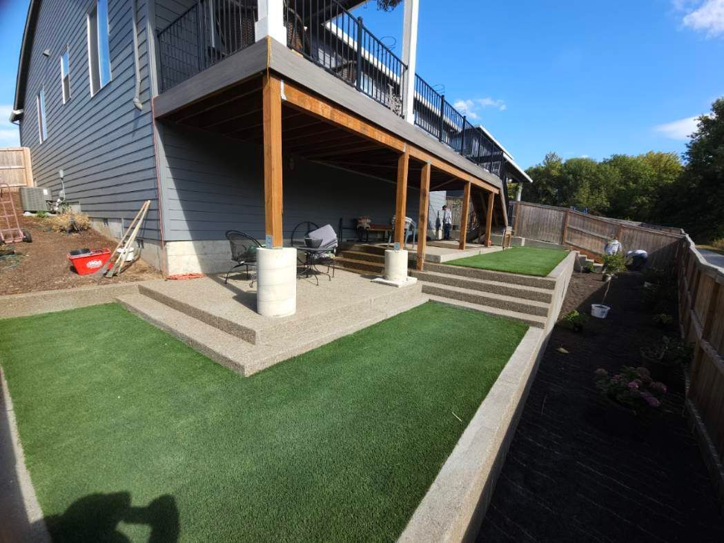 Backyard patio with raised deck, steps, and green turf beside a house and retaining wall