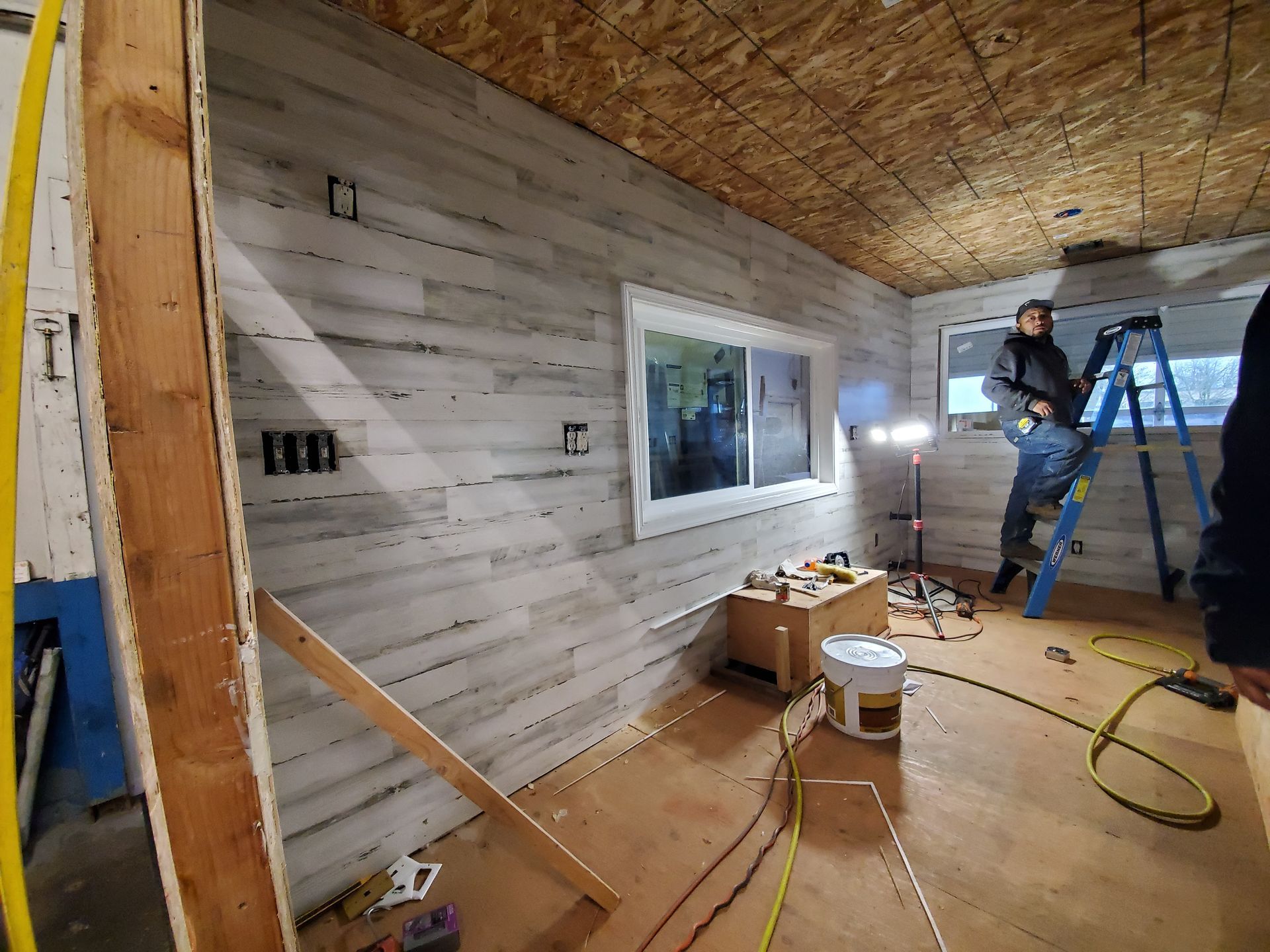 Worker installing white shiplap wall in a room under construction, with tools and ladder nearby.