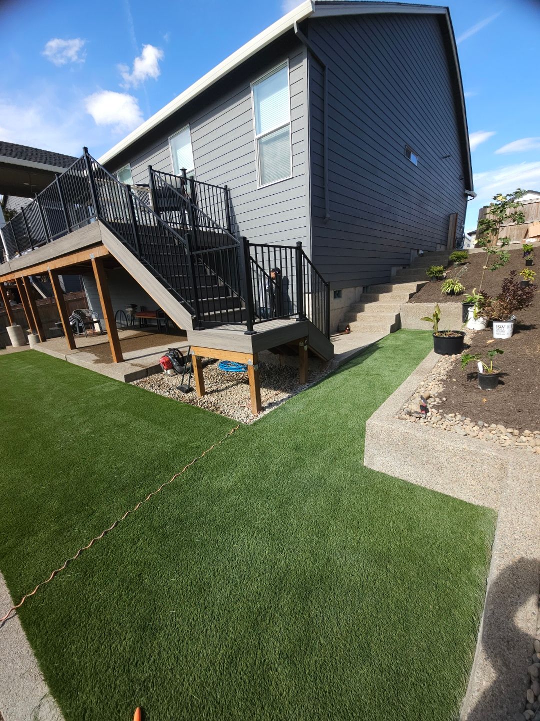 Backyard view of a gray house with black stairs, patio, and artificial grass under a blue sky