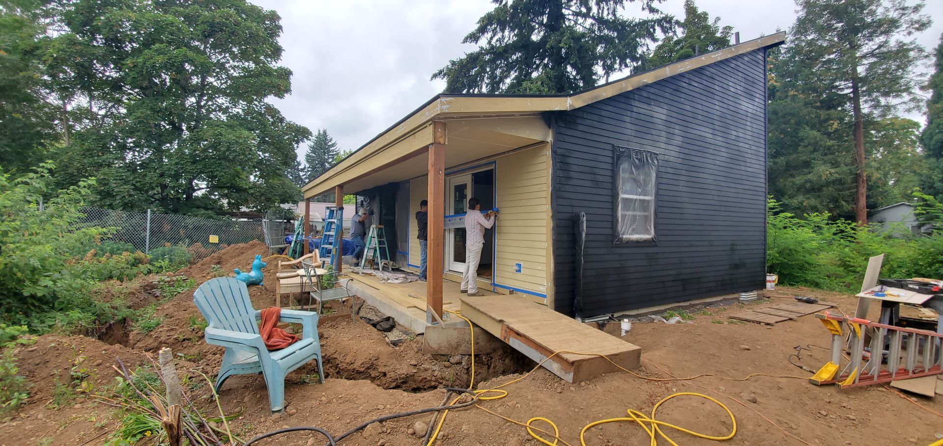 Partially renovated house exterior with black siding, porch posts, and construction debris in a wooded yard