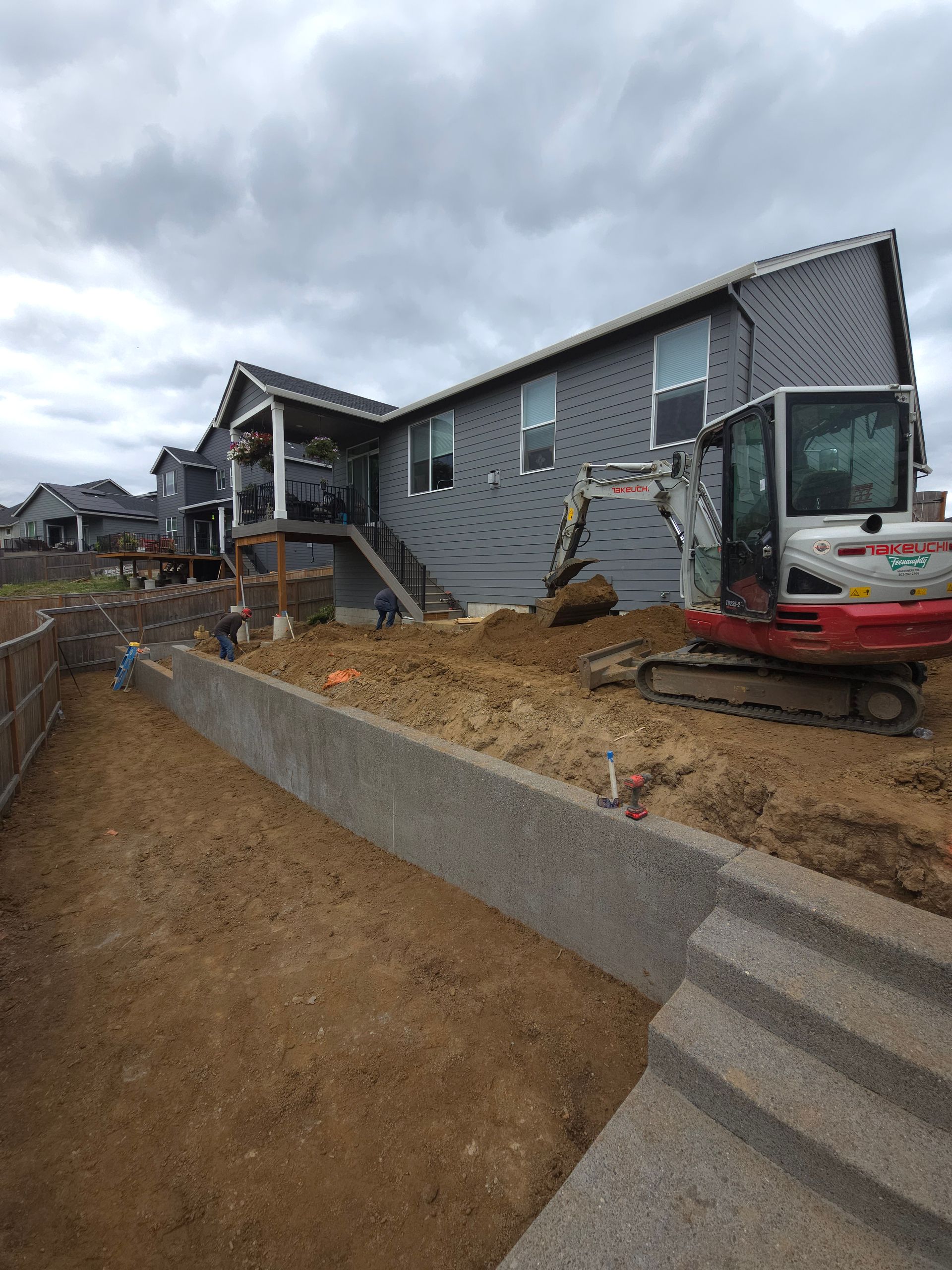 House under construction with excavator, dirt retaining wall, and cloudy sky