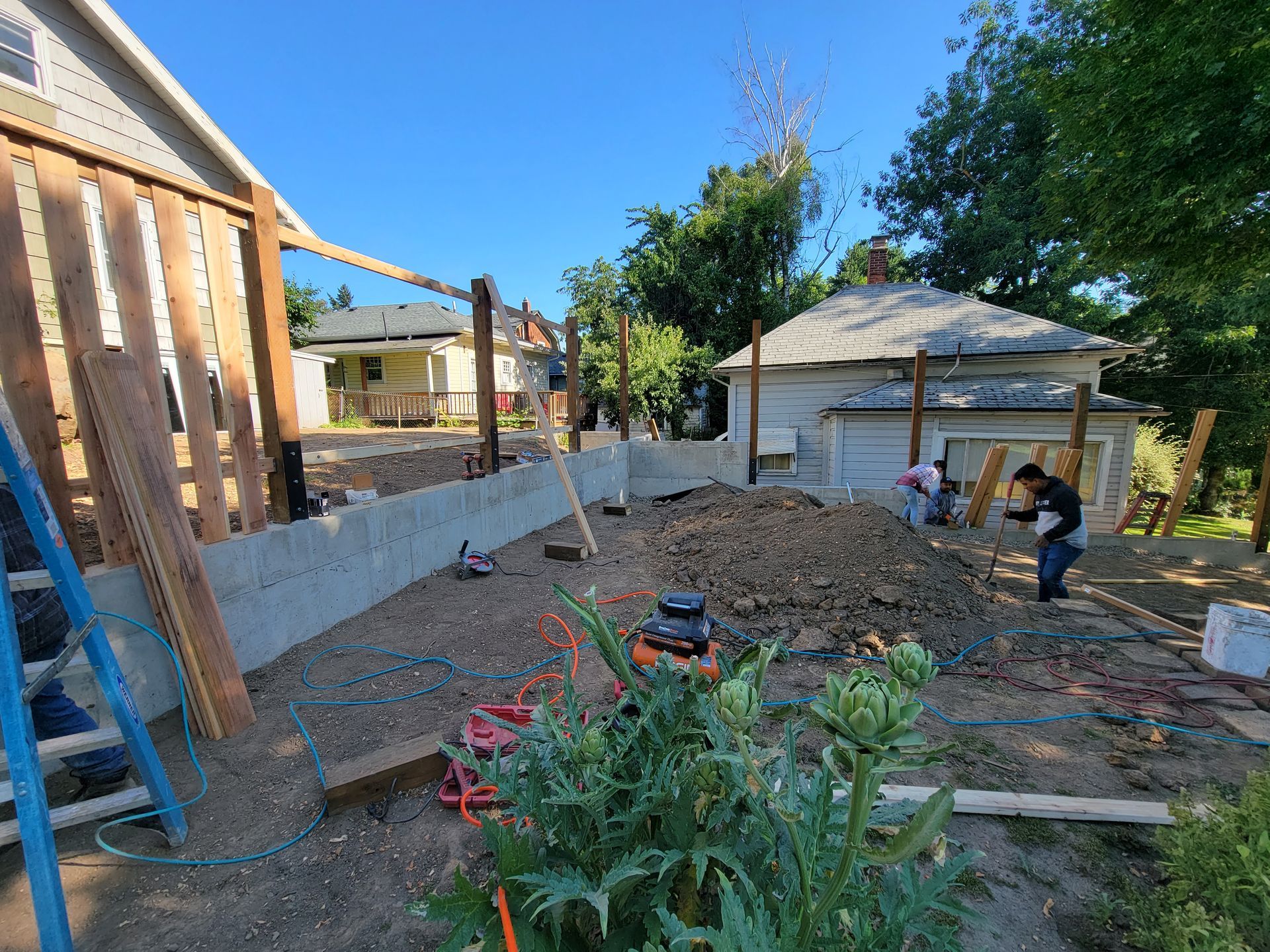 Backyard construction site with dirt mound, workers, wooden fence, and a house in the background