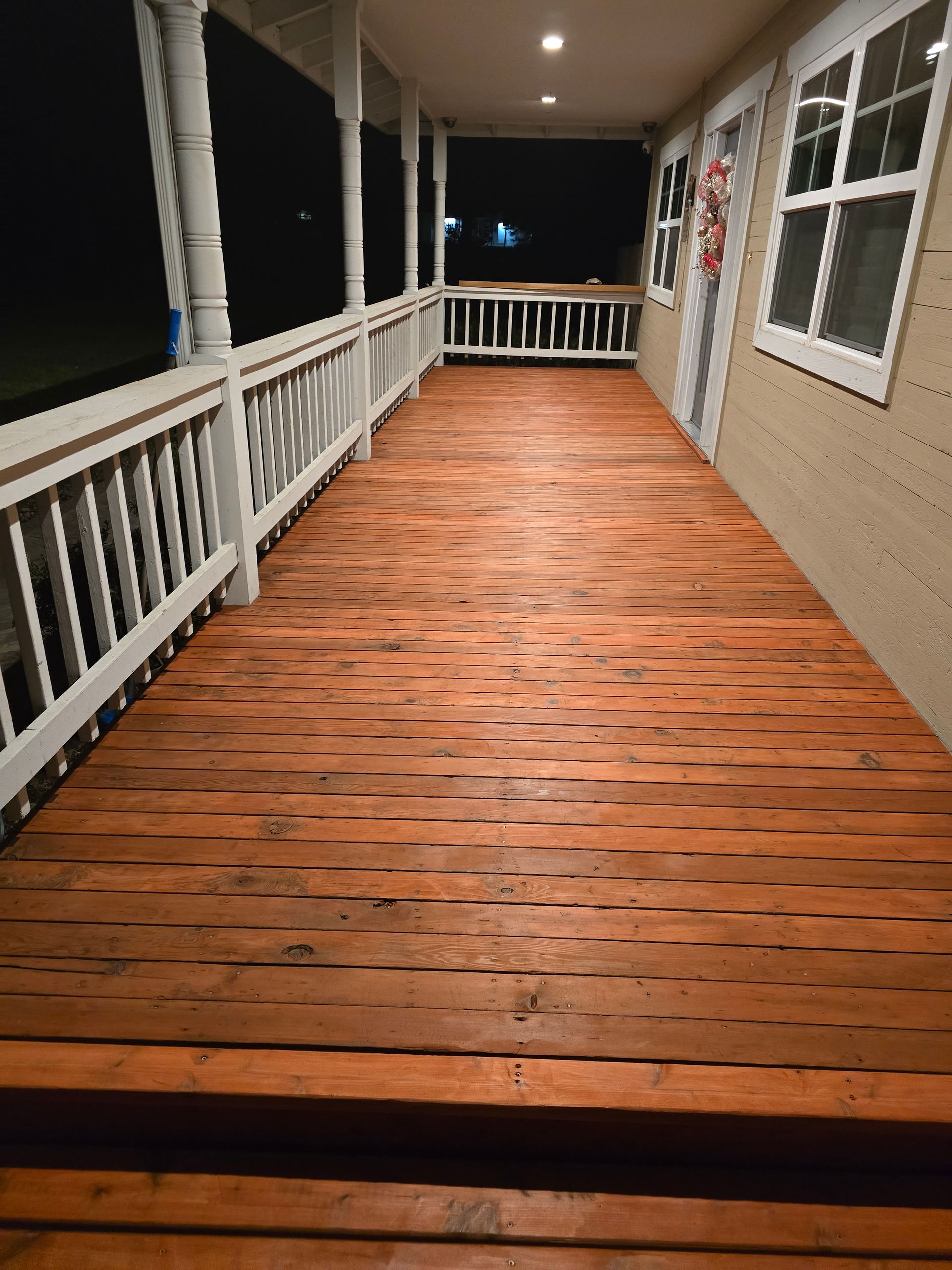 Wooden porch at night with white railings, beige siding, and brick-like floorboards under warm ceiling lights
