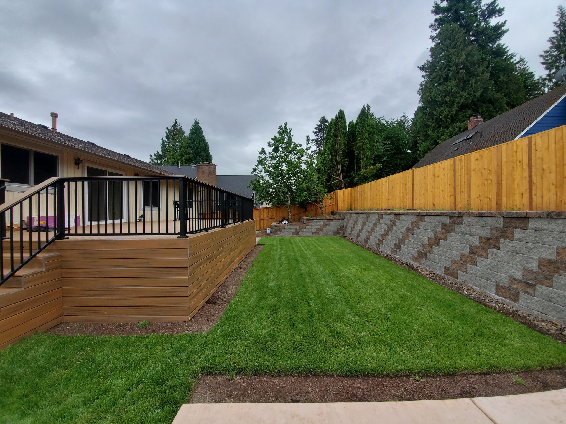 Backyard with sloped grass lawn, wooden deck, and fenced retaining wall under cloudy sky