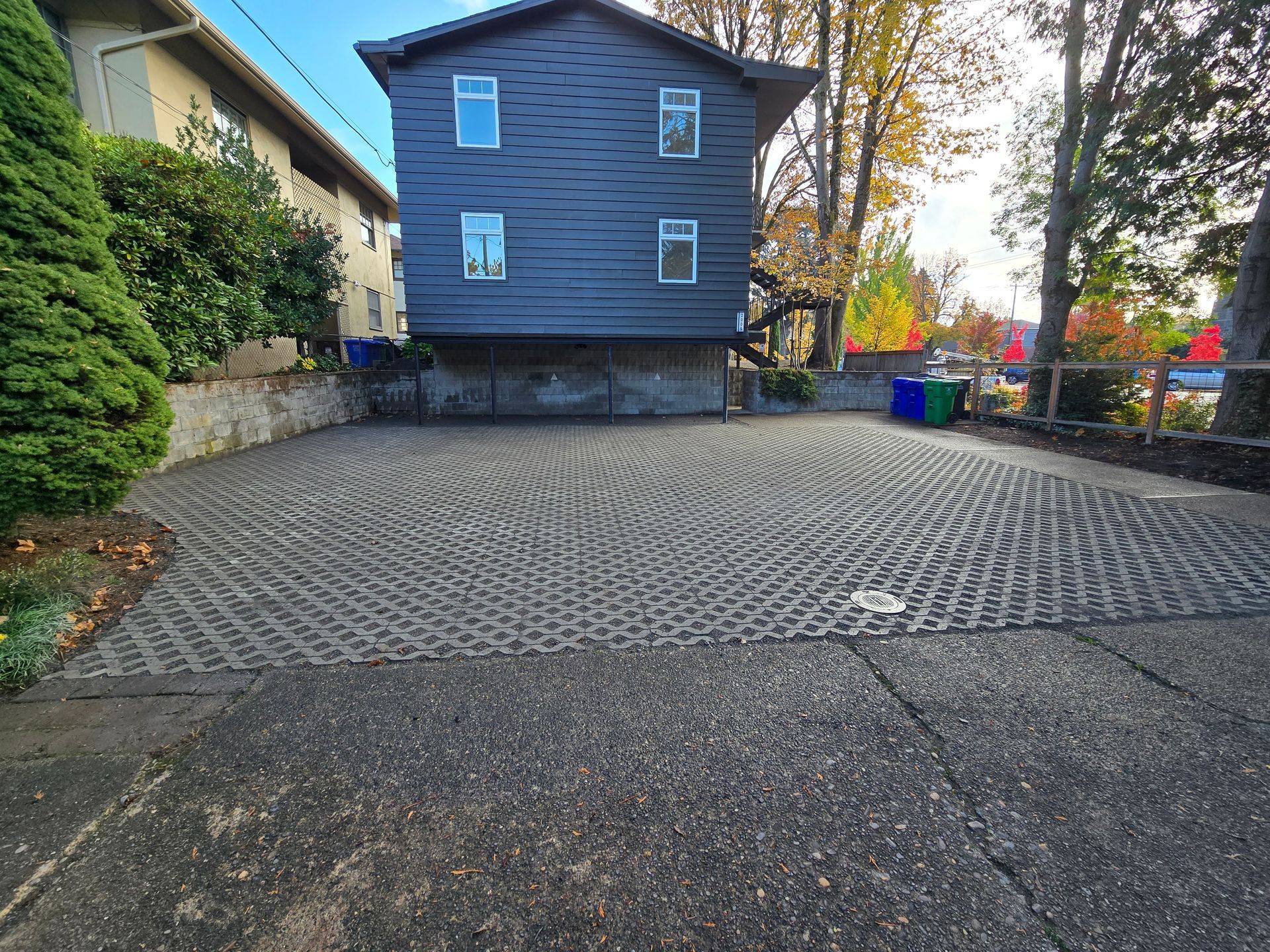 Cobbled parking lot beside a dark wooden house with trees and bins in the background