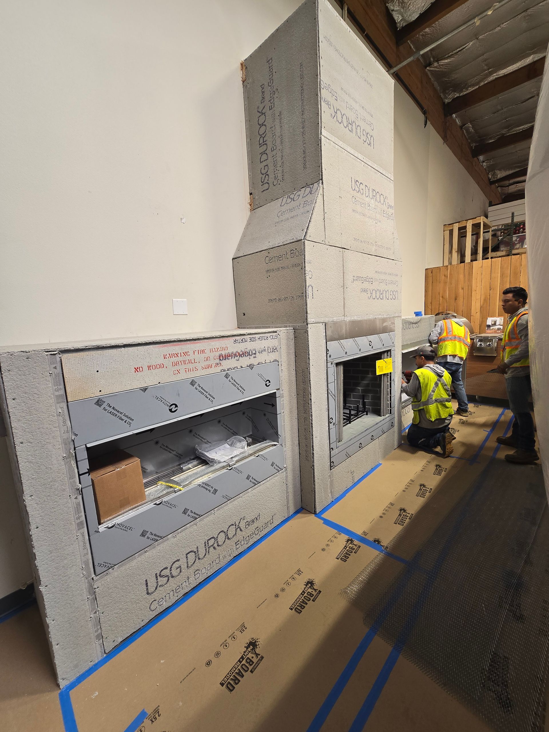 Construction workers installing wall panels in a narrow interior corridor, with cardboard floor protection and blue tape.