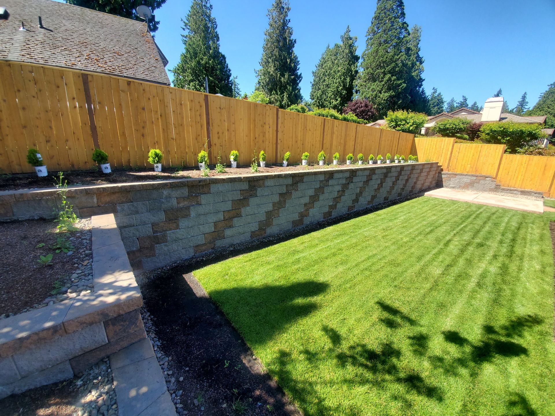 Backyard with green lawn, stone retaining wall, wooden fence, and potted plants under a blue sky